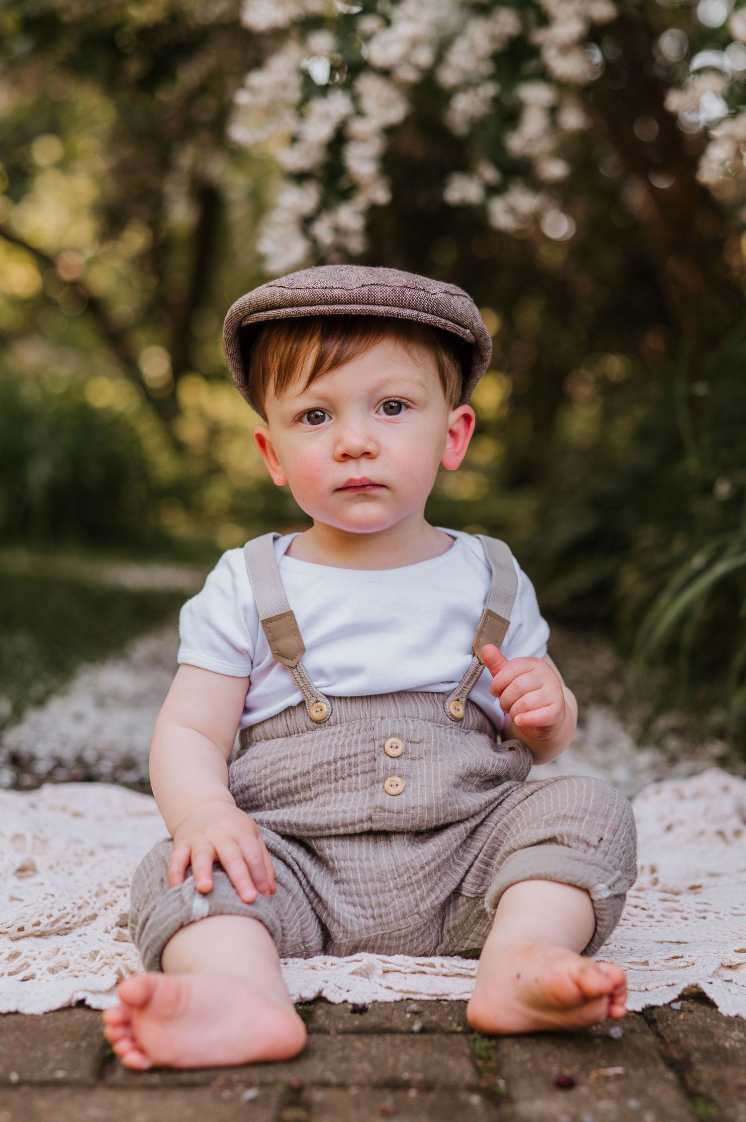 Baby wearing brown flat cap and gray overalls sits on a blanket in a garden setting with soft bokeh background.