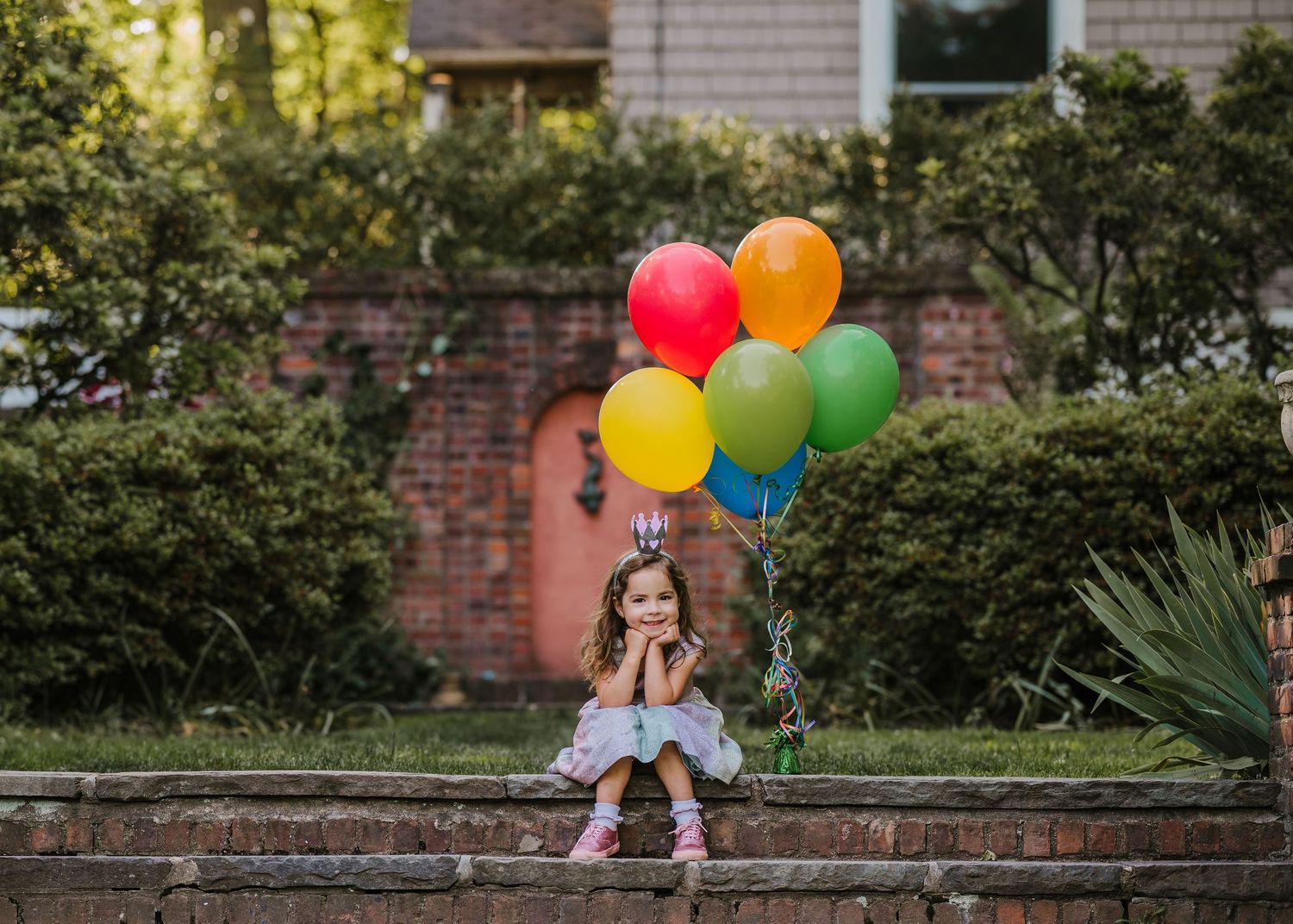 Colorful rainbow balloons add a cheerful touch to a garden scene with brick steps and shrubbery.