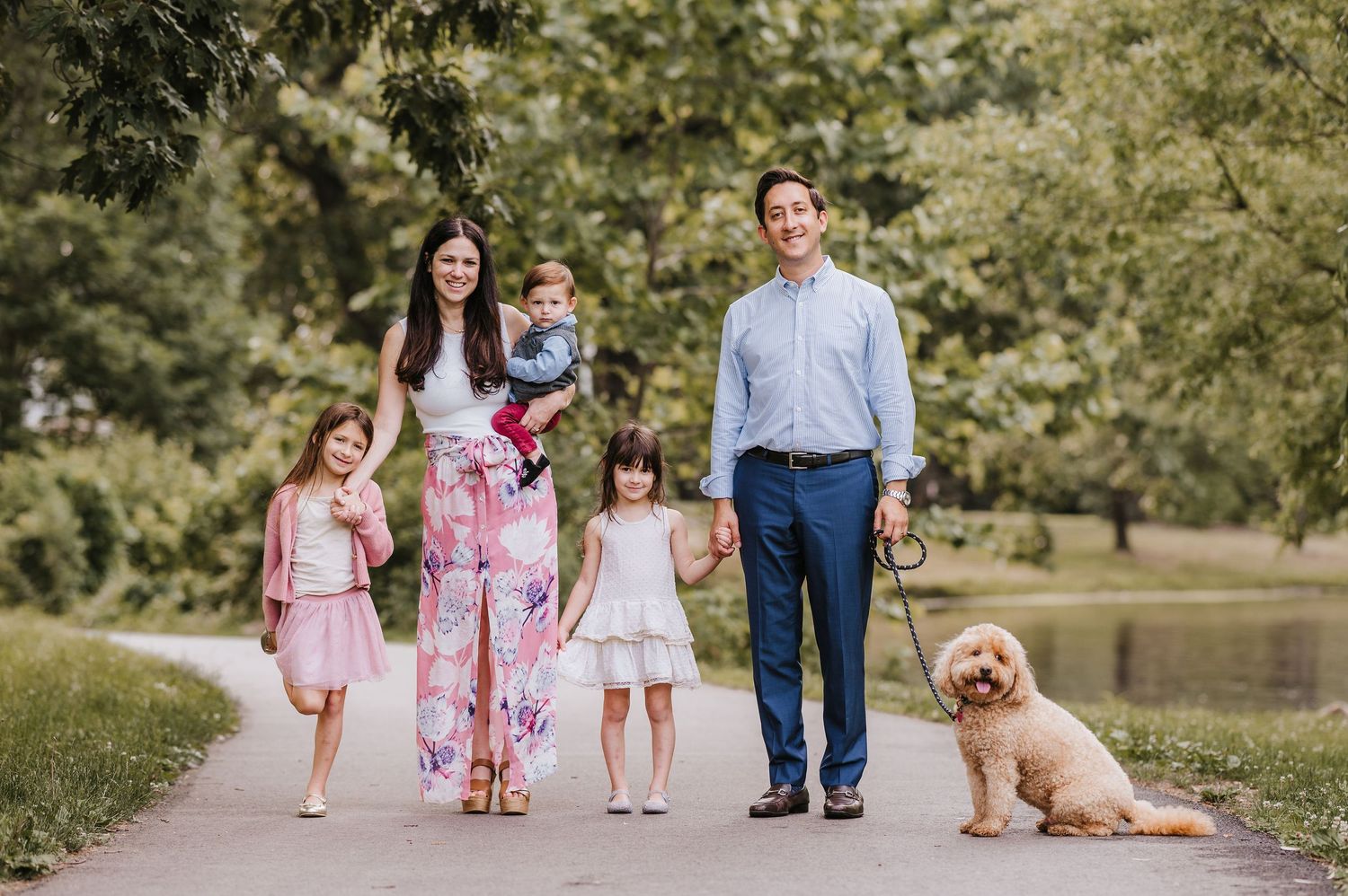 A family walking together on a park path with their golden retriever dog while surrounded by lush green trees.
