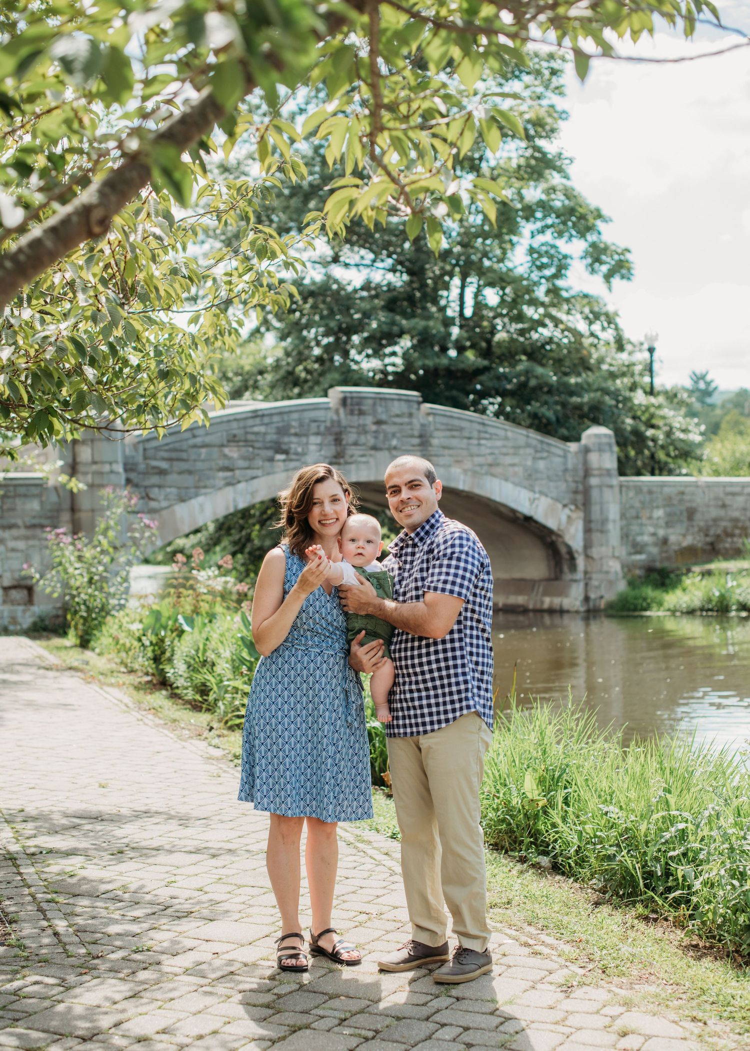 A couple holding their dog poses together on a towpath beside a stone bridge and canal on a sunny summer day.