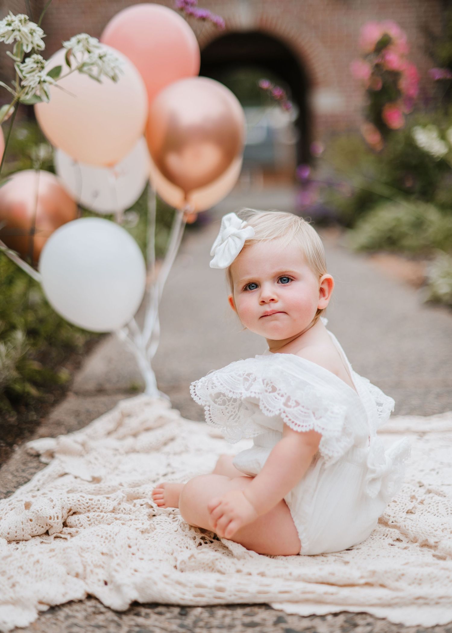 Baby in white dress sitting on lace blanket with pink and white balloons in garden setting.