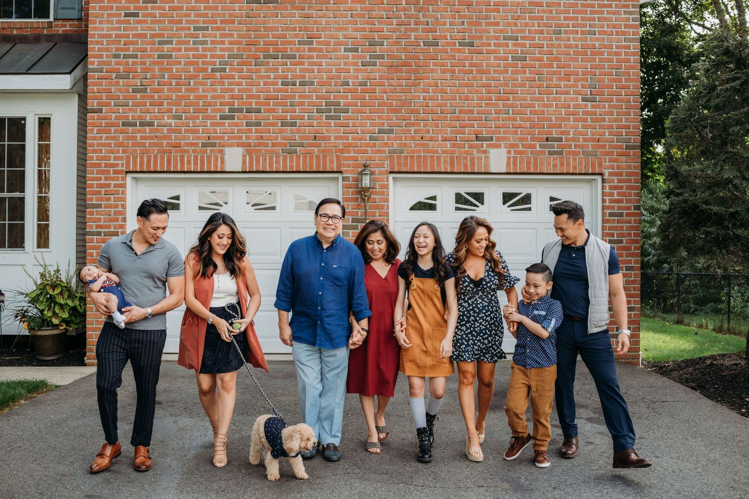 A group of young adults and a small dog stand together in casual attire in front of a brick house garage.