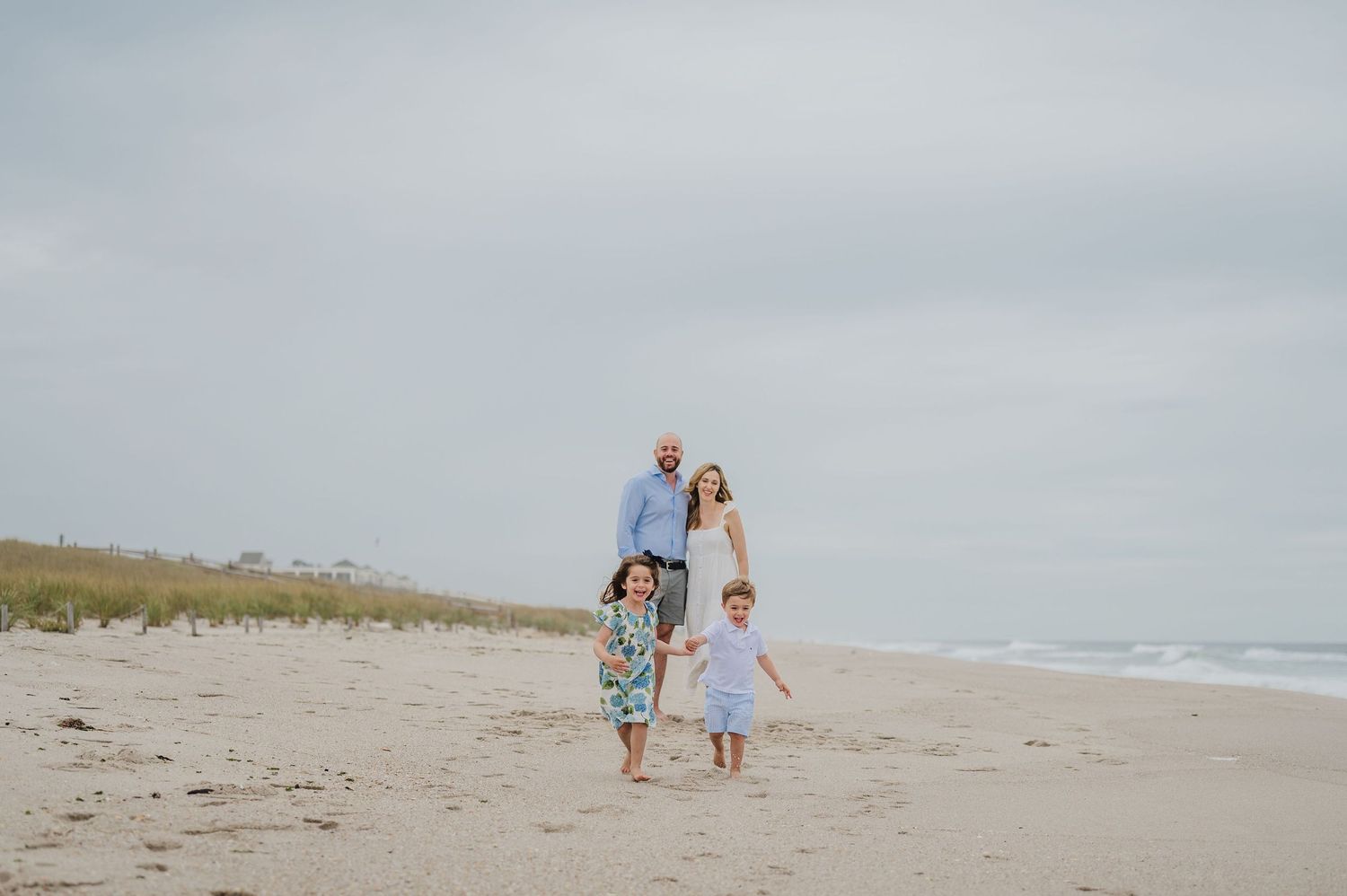 A couple walks hand in hand on a cloudy beach with waves crashing in the background as their children run ahead.