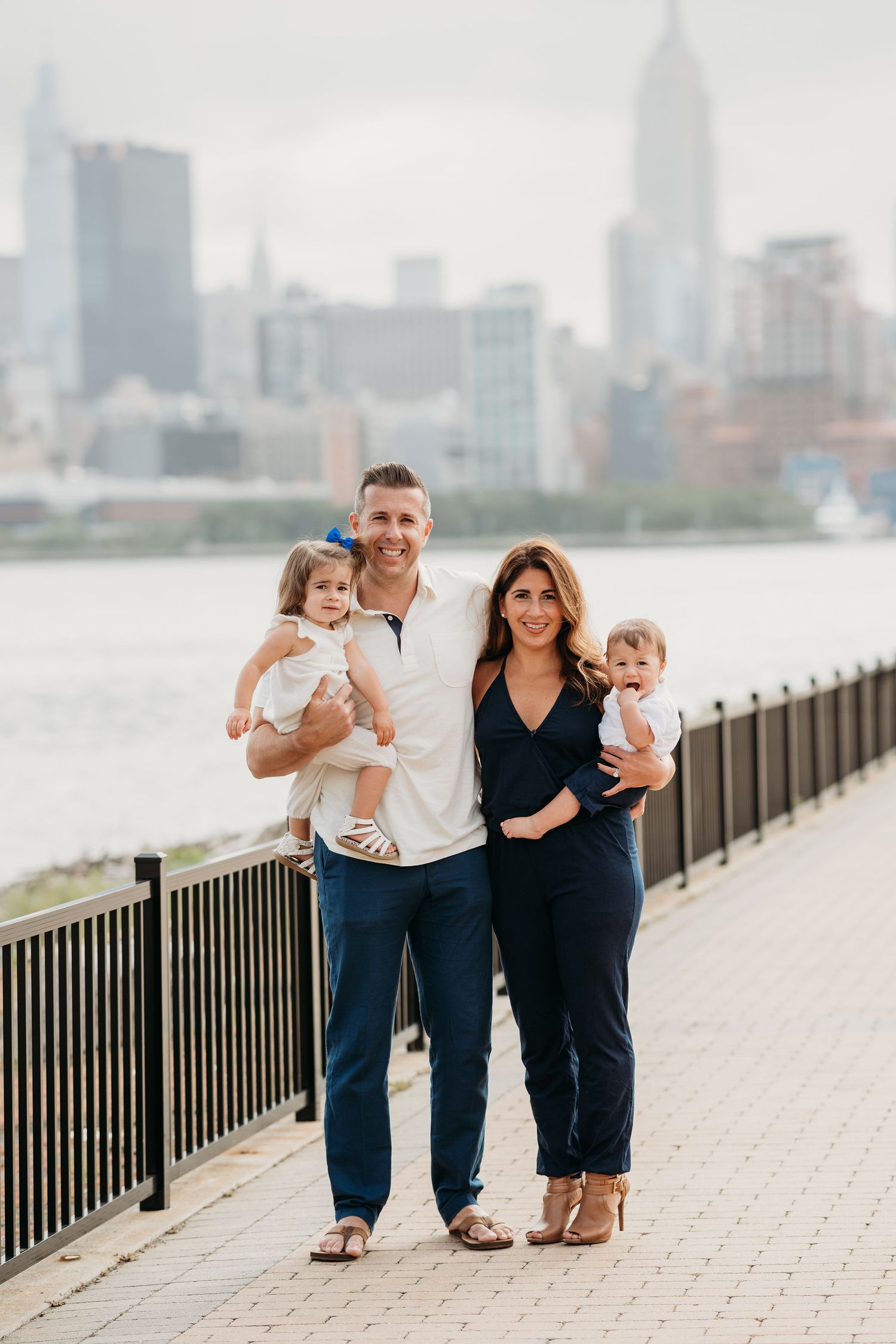 Family stands together along waterfront promenade with NYC skyline visible in background on overcast day in Hoboken.