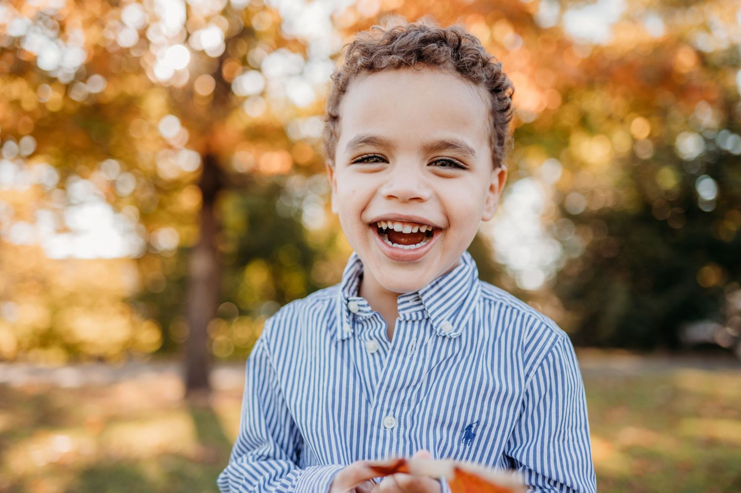 A cheerful young person in a blue striped button-down shirt smiles outdoors against fall foliage in Summit and Millburn,