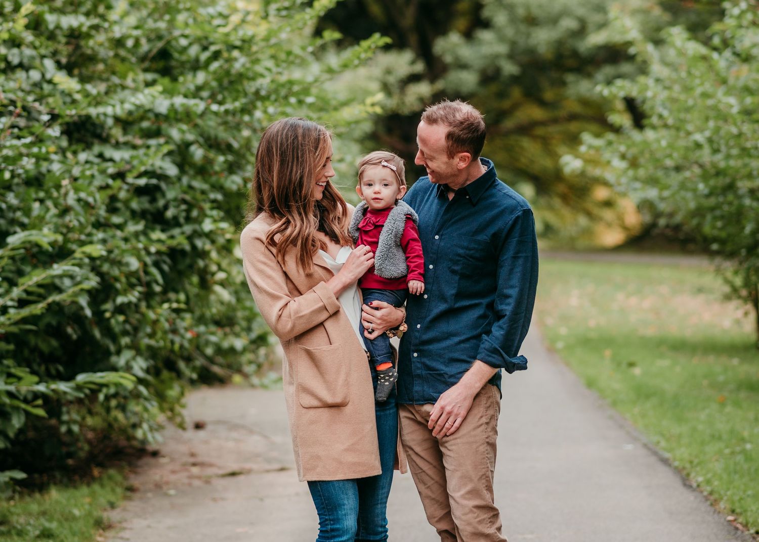A couple in autumn clothing walks together with their young child on a garden path surrounded by lush greenery.