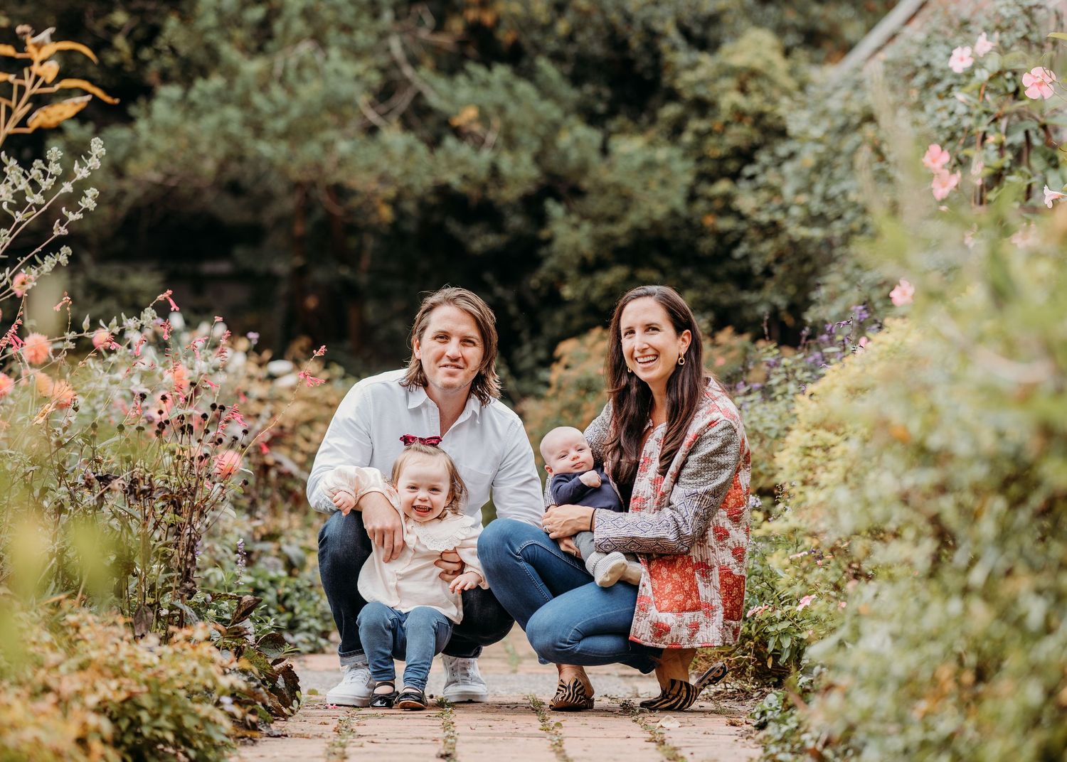A family kneels together on a garden path surrounded by blooming flowers and green foliage in a natural setting.