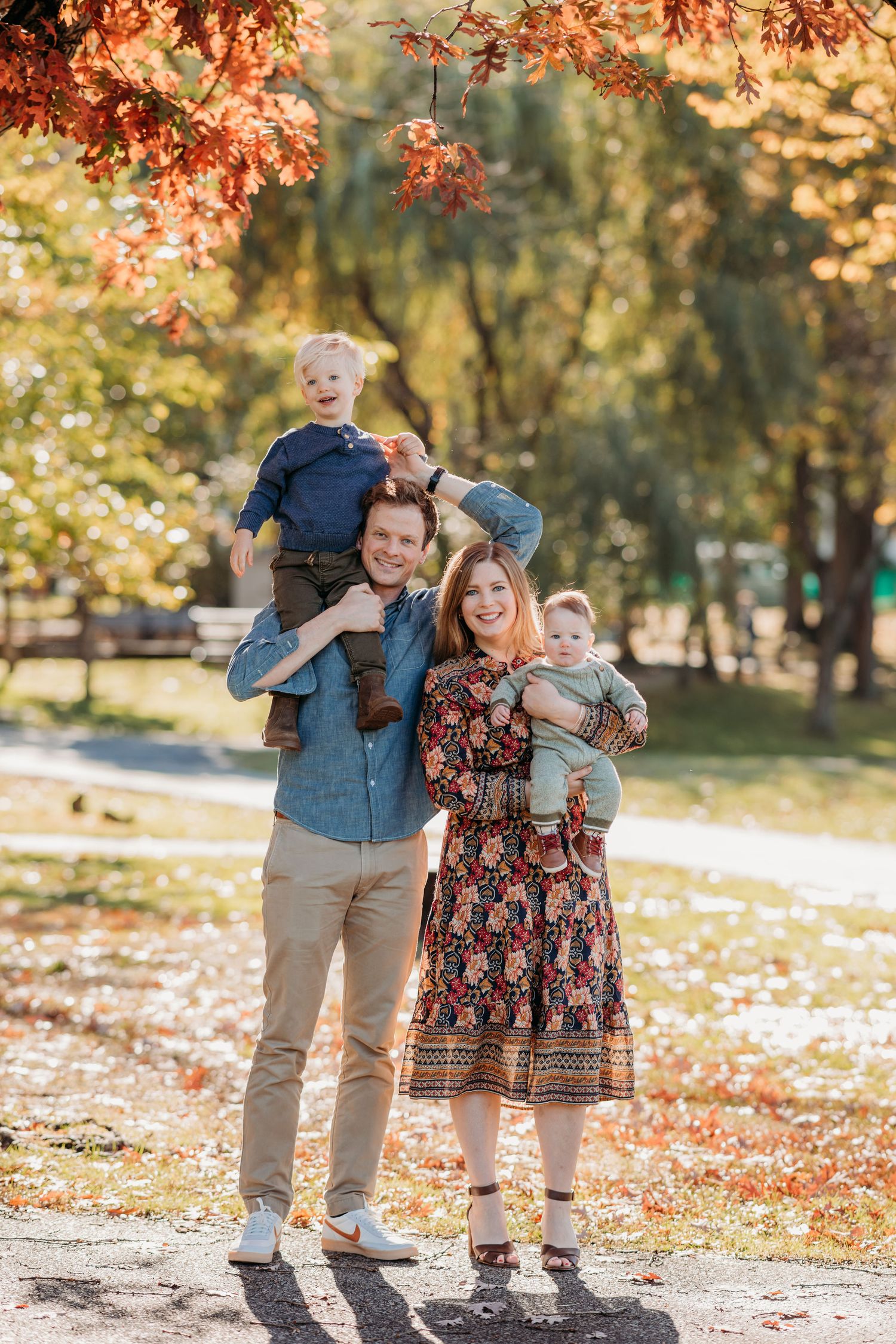 A family stands together on an autumn day with fall foliage and golden sunlight in a park setting.