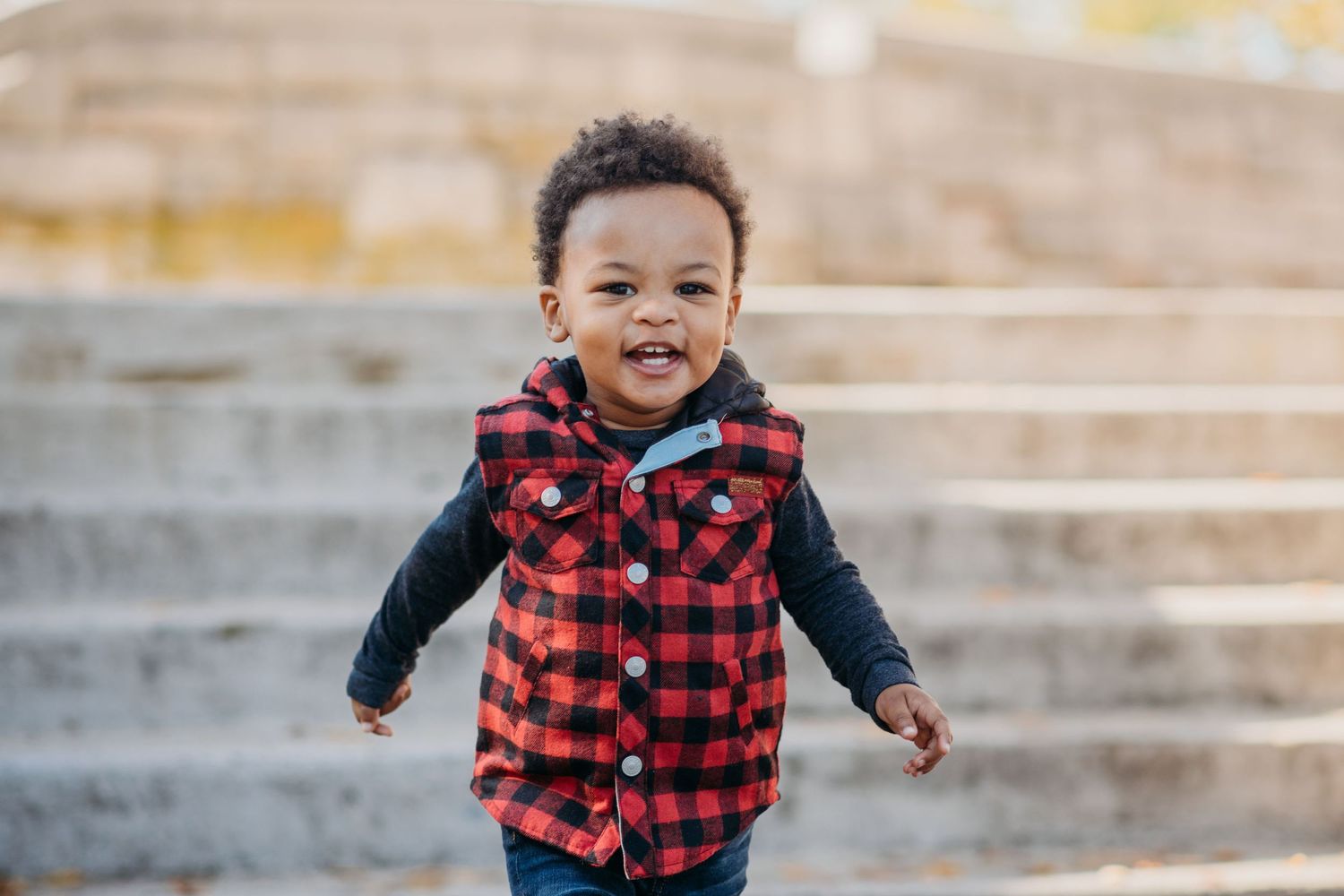 Happy toddler wearing red plaid vest and long sleeve shirt running on outdoor steps on autumn day in Verona.