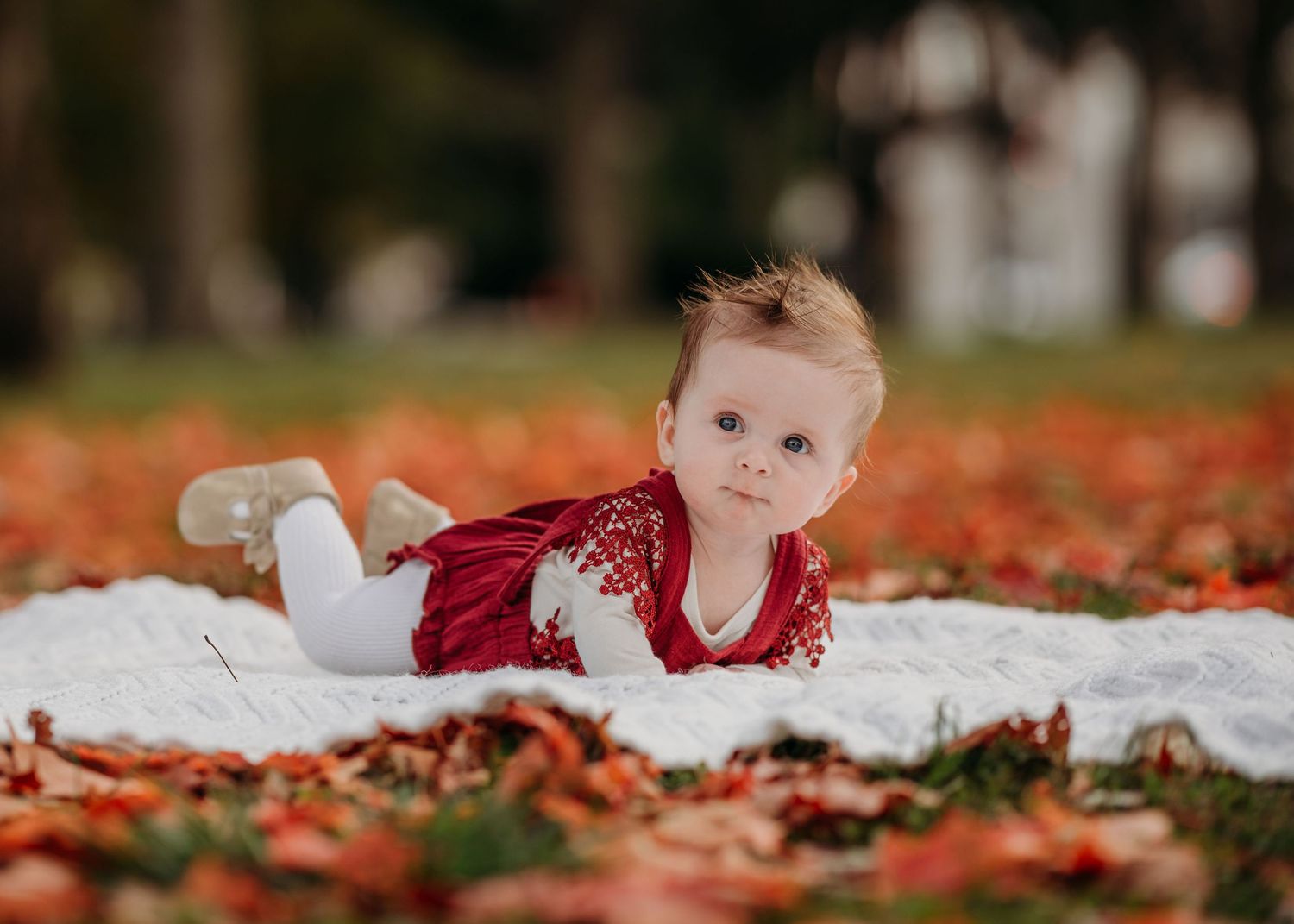 Baby in red velvet dress lying on white blanket surrounded by fall leaves in an autumn outdoor setting.