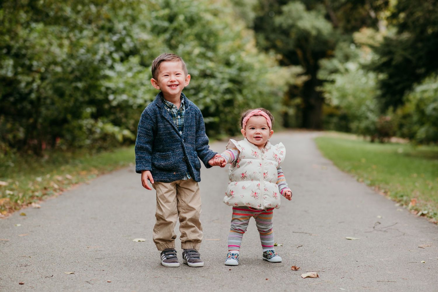 Two young children holding hands and laughing while walking together on a tree-lined path.