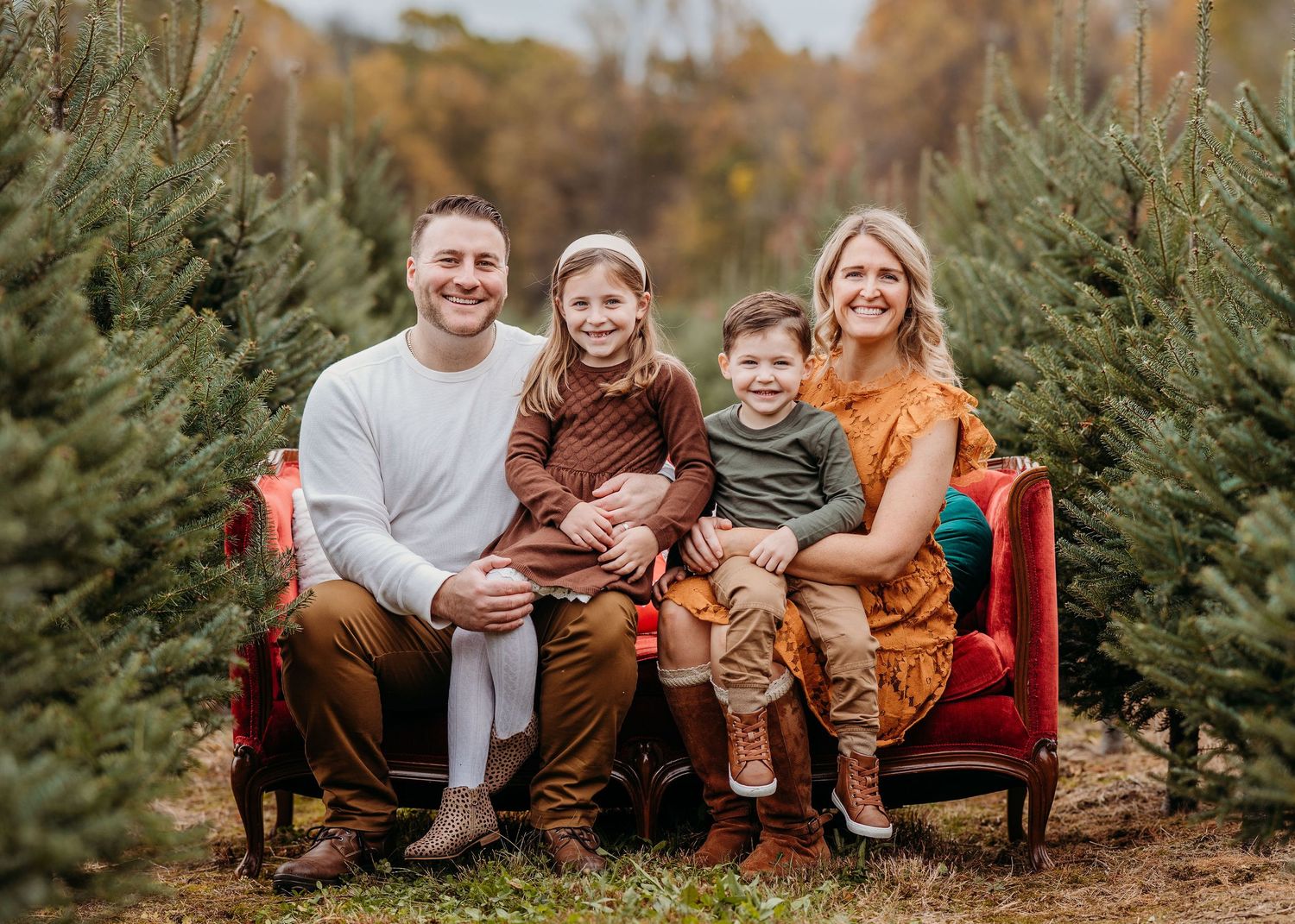 A family sits on a red bench amongst evergreen trees at a Christmas tree farm during autumn.