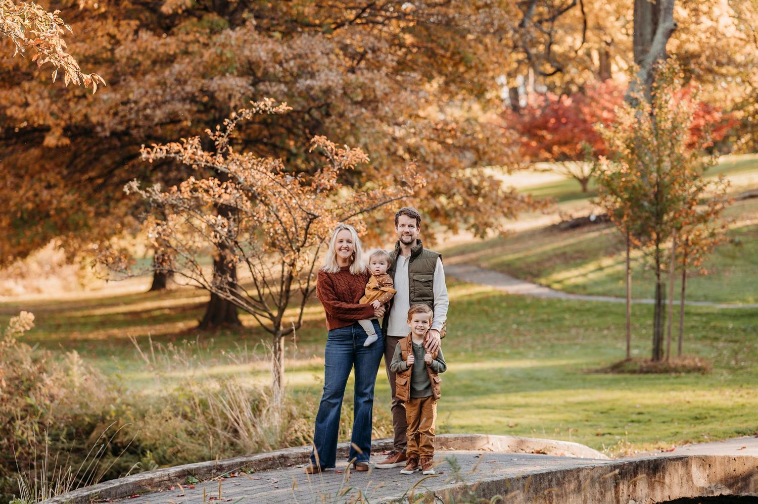 A couple and their dog stand together on a path surrounded by autumn foliage in a scenic park setting in New Jersey.