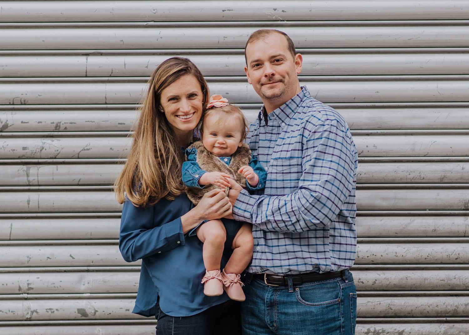 A family poses together against a corrugated metal wall background wearing blue and plaid clothing in Hoboken.