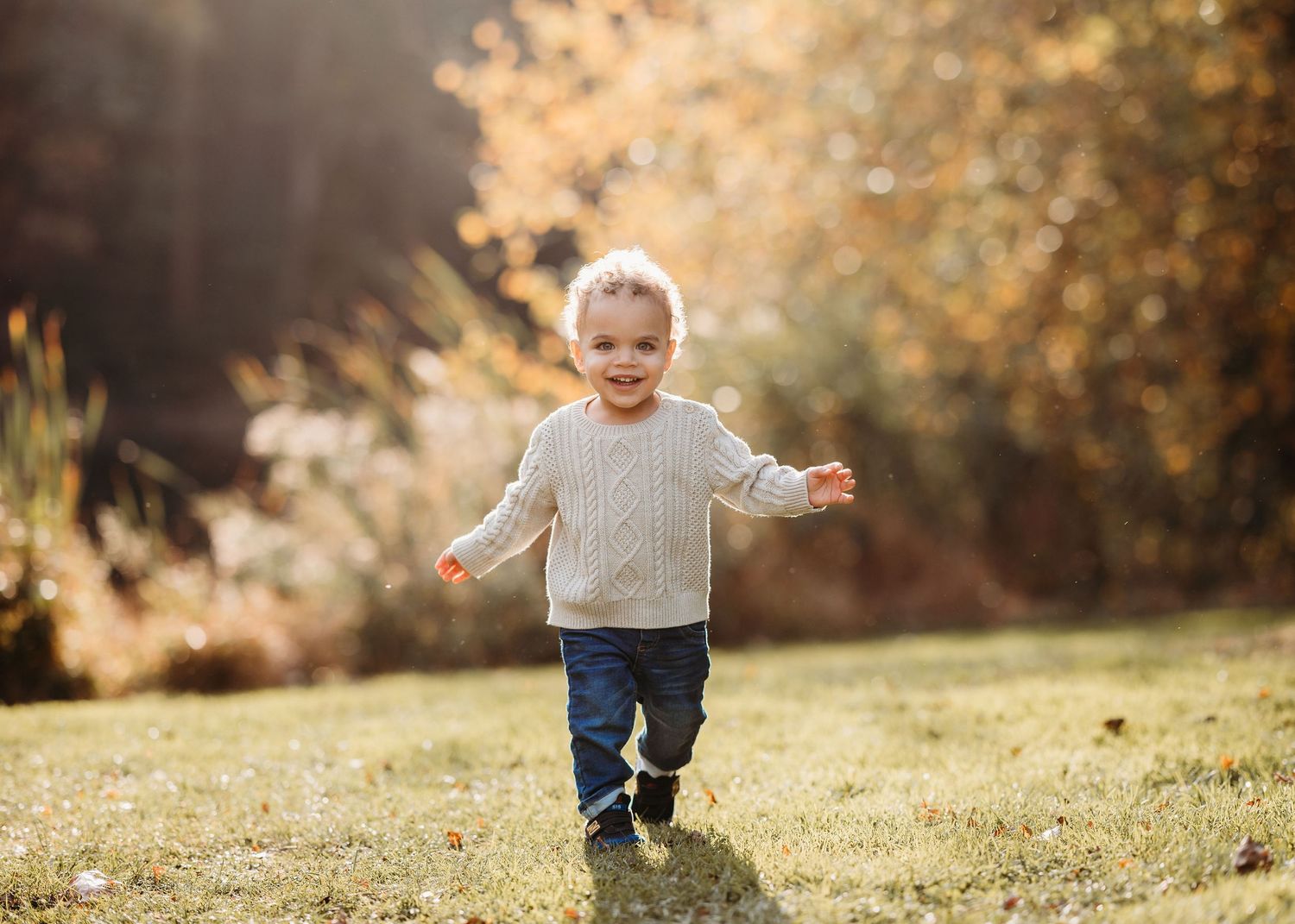 Child running joyfully through a sunny autumn meadow wearing a creamy sweater and jeans in Millburn.