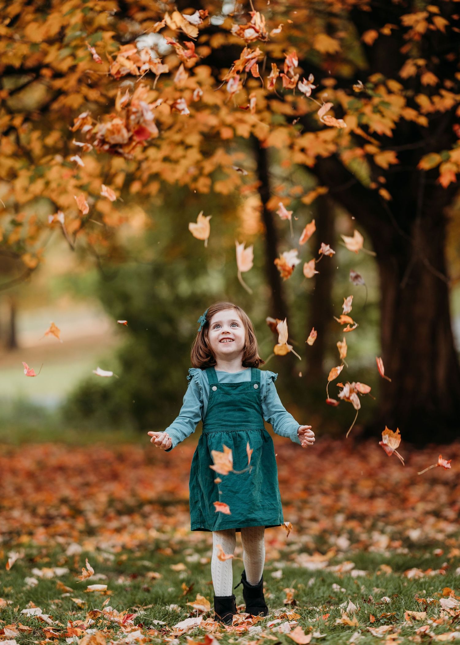 Autumn leaves fall around a small figure in a green dress playing joyfully in a park during golden hour.