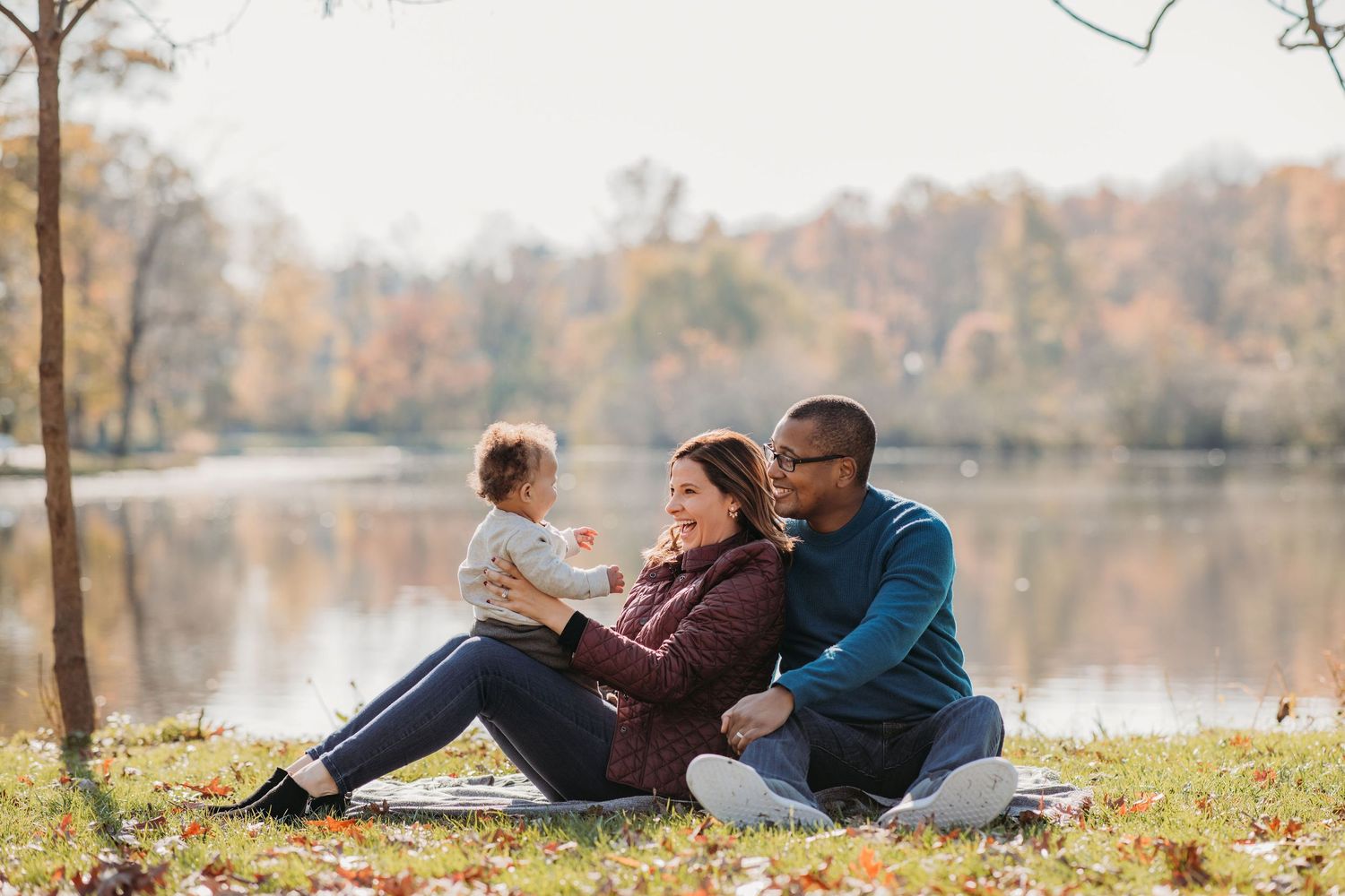 Family enjoying autumn day sitting together on blanket by serene lake with fall foliage in background.