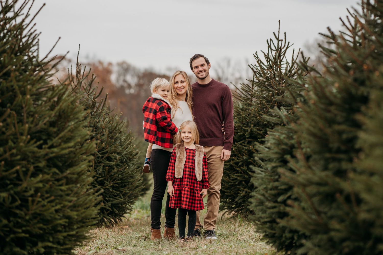 A family stands together between rows of evergreen trees at a Christmas tree farm on an autumn day.