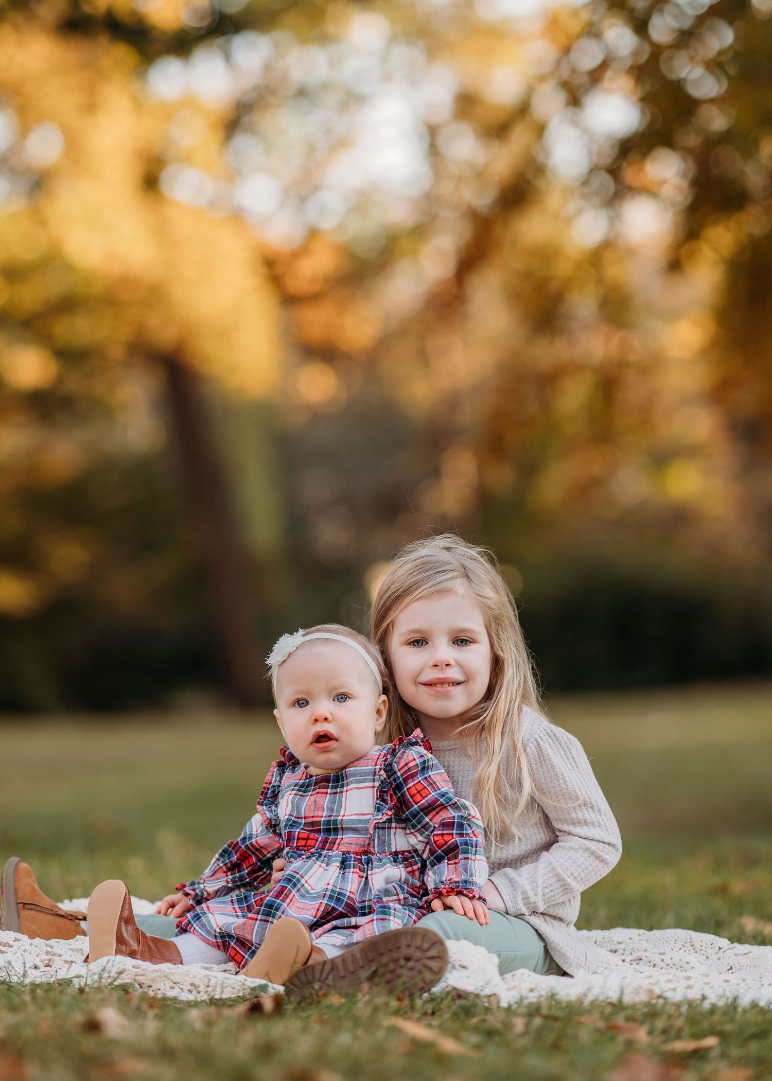 Two children sit on a white blanket outdoors during autumn with golden foliage bokeh in the background.