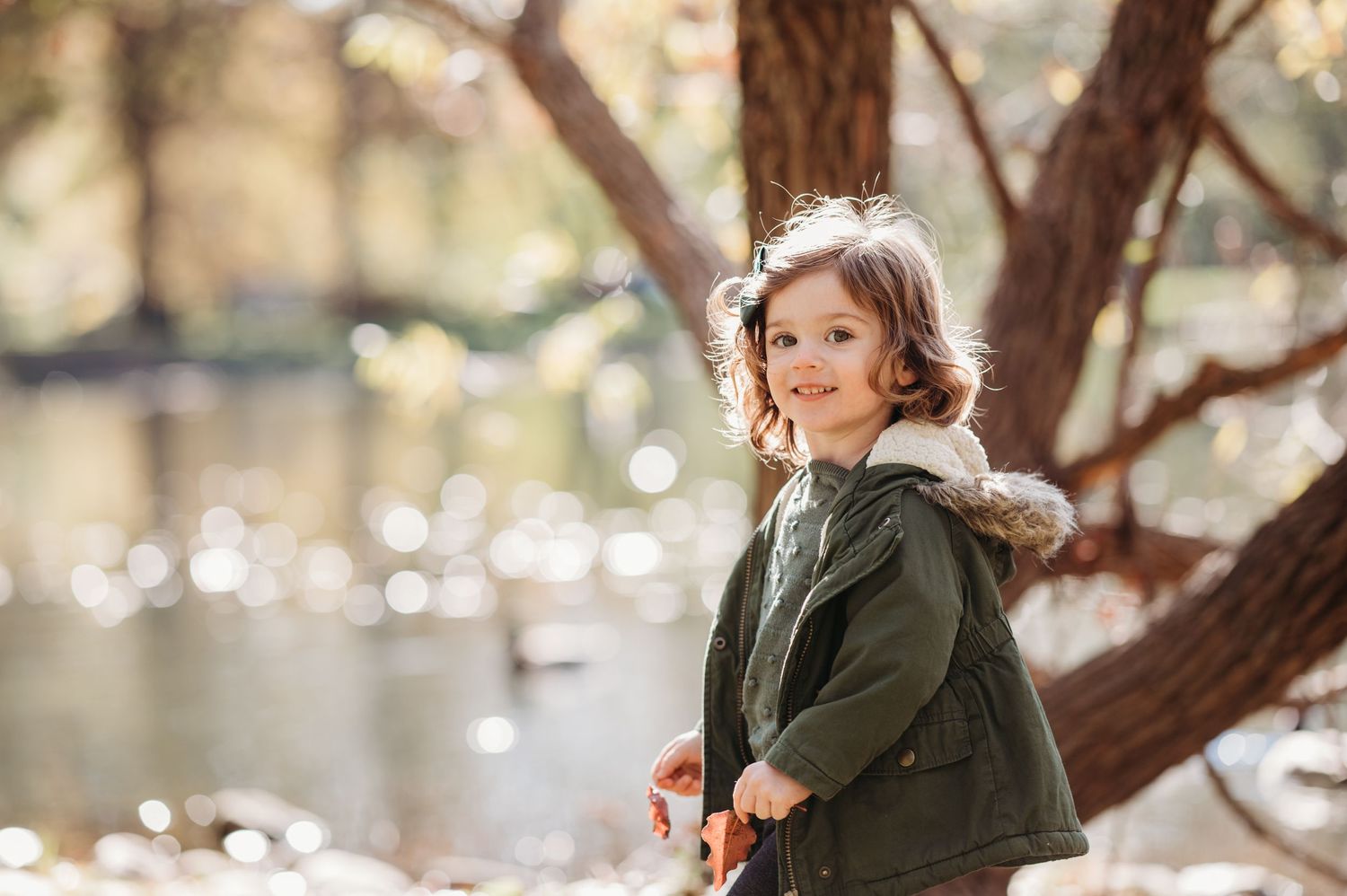 Child in green winter coat stands beside tree trunk with bokeh lights in background.