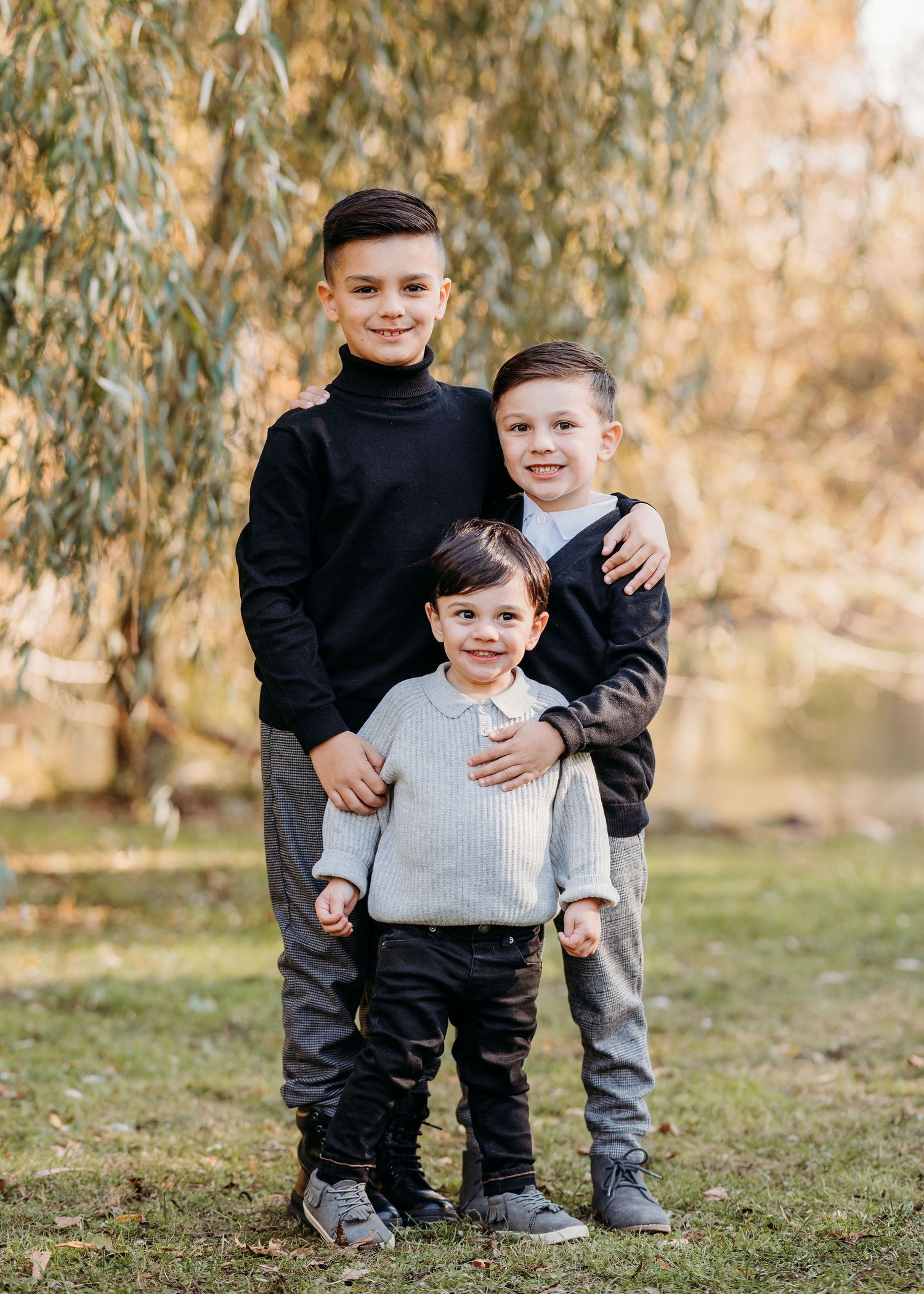 Three young children in casual outfits posing together outdoors with a willow tree in the background.