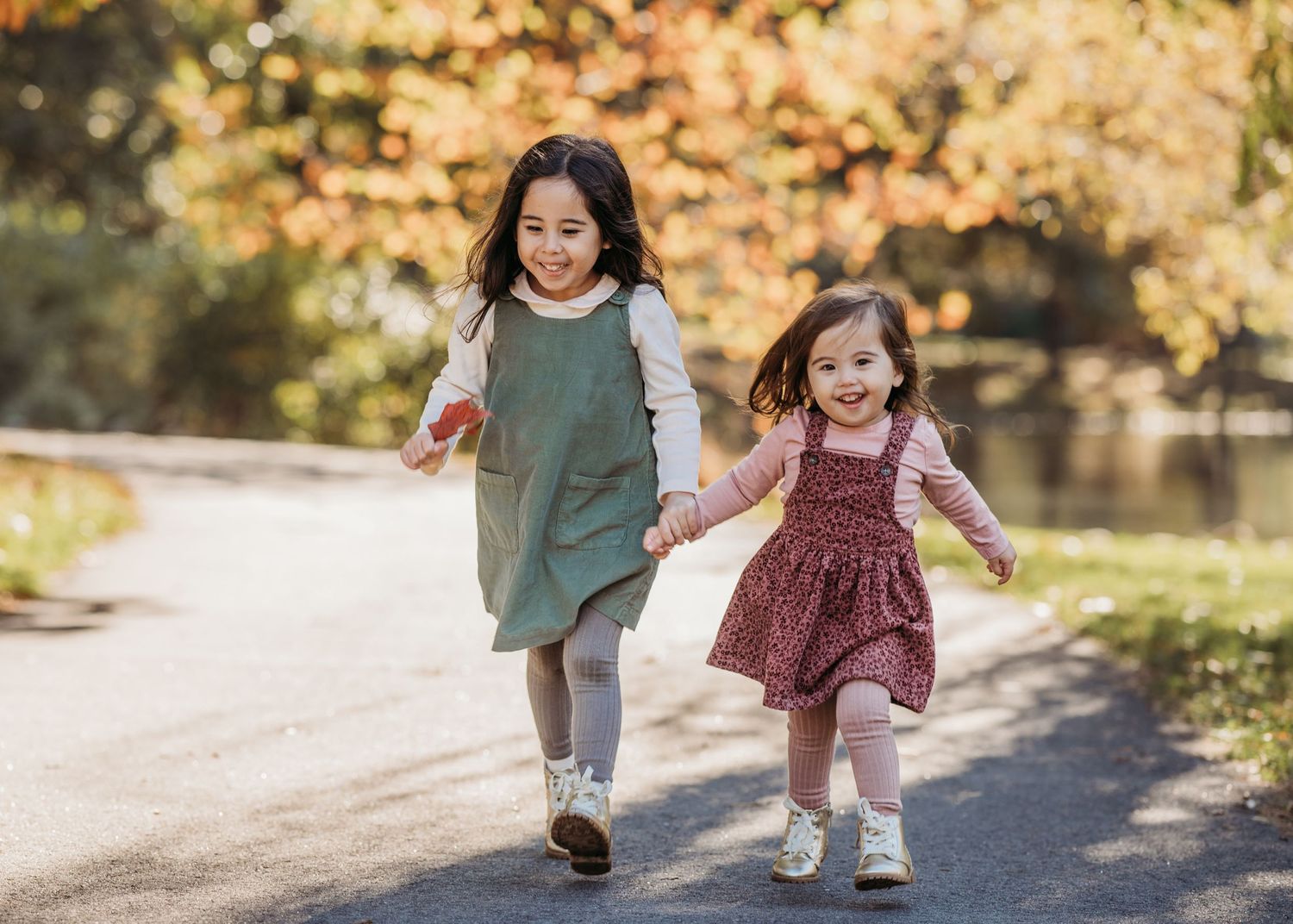 Two young children run hand-in-hand down a sunny path surrounded by autumn foliage and a pond.
