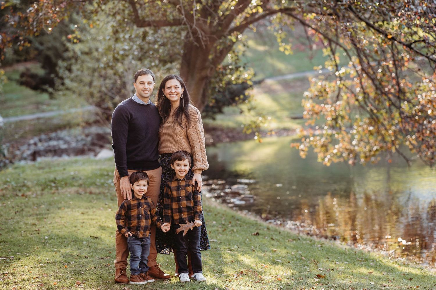A family stands together next to a lake in autumn, wearing coordinating plaid and dark sweaters in a natural outdoor setting.