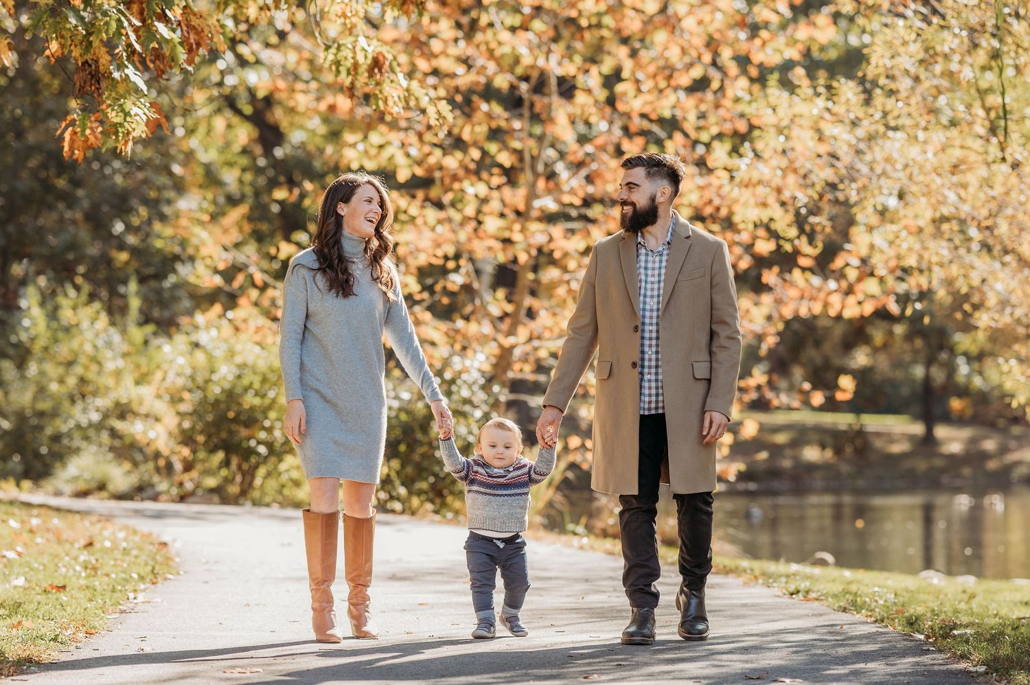 Family walks together holding hands on autumn path near lake with golden fall foliage in background.