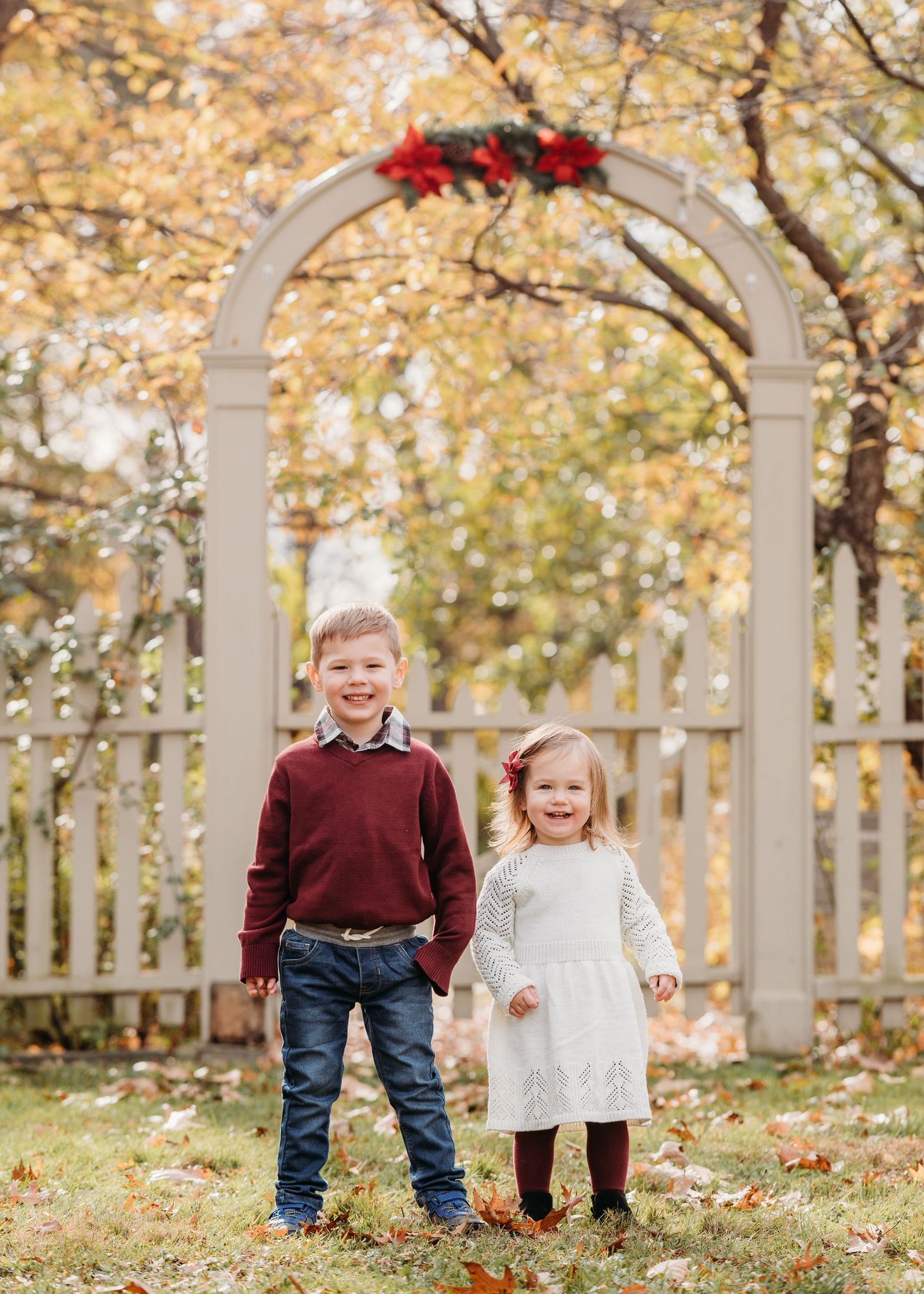 Two young children stand beneath a white wooden arbor decorated with red flowers on a sunny autumn day in Bloomfield.