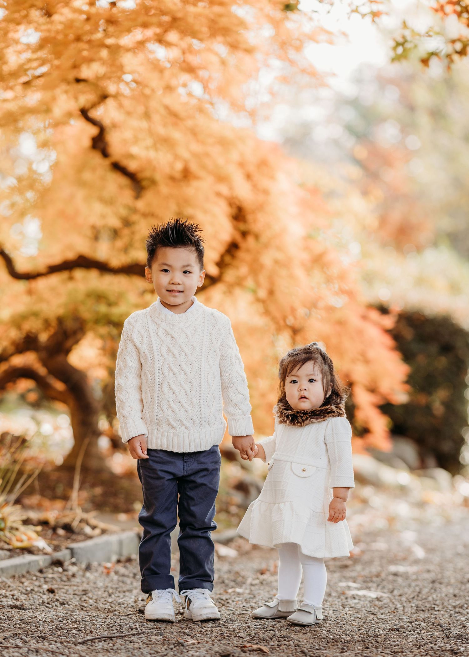 Two small children holding hands stand together in front of vibrant orange autumn foliage on a dirt path in Montclair.