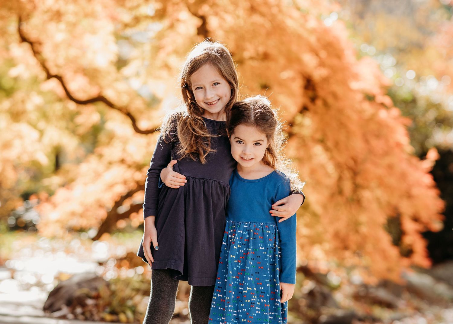 Two children in navy blue dresses stand together outdoors with vibrant autumn foliage in the background.