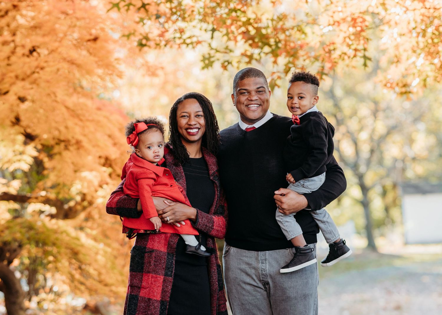 Family of four poses together outdoors during autumn with vibrant orange fall foliage in the background.