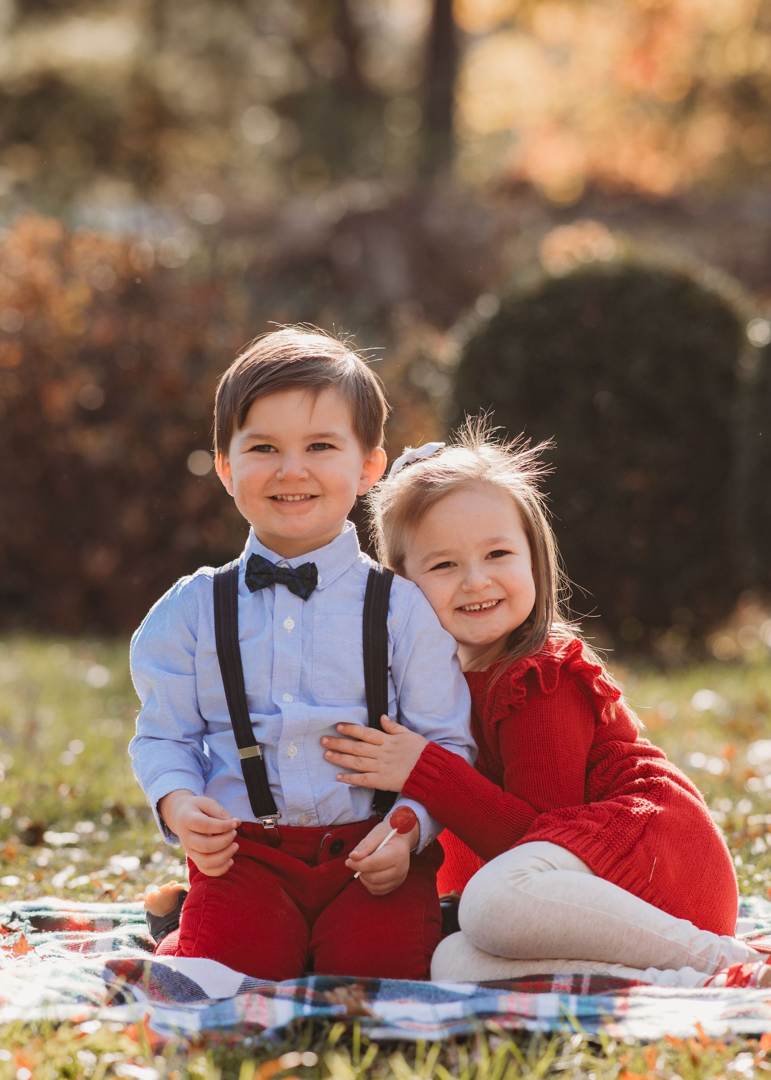 Two children in formal attire sit together on a blanket during autumn with fallen leaves in the background.