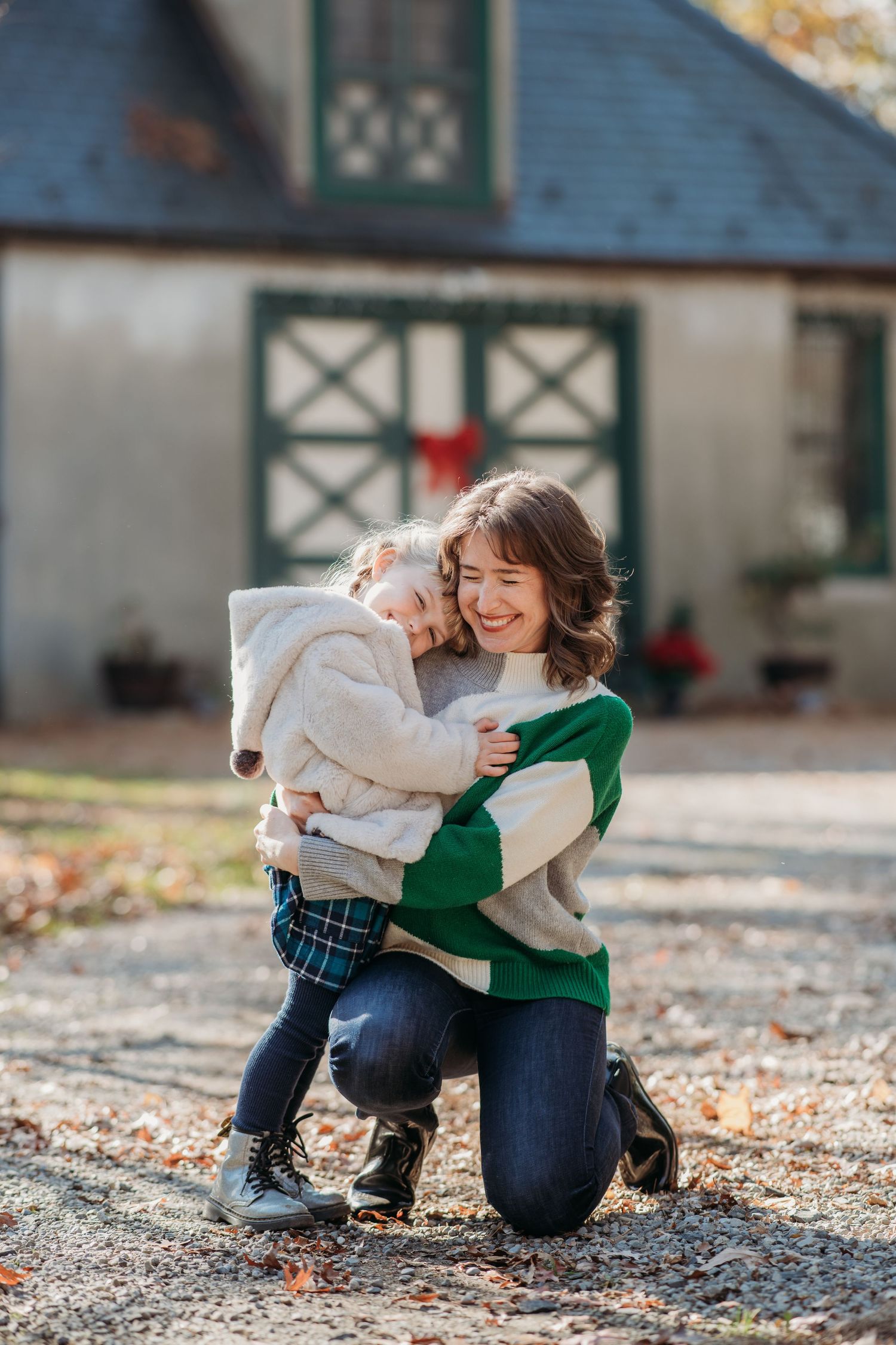 A parent and child share a joyful embrace outdoors in autumn, kneeling on fallen leaves near a rustic building.