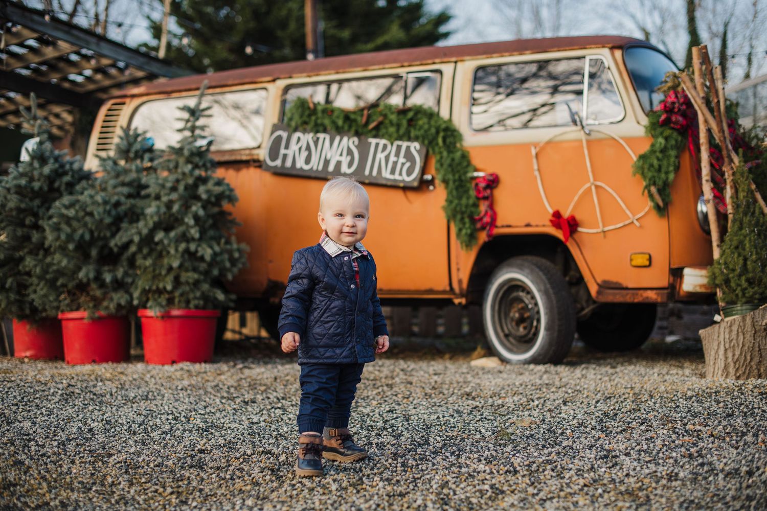 A small child stands in gravel near an orange vintage VW van with holiday decorations and greenery outside at La Serra in totowa.