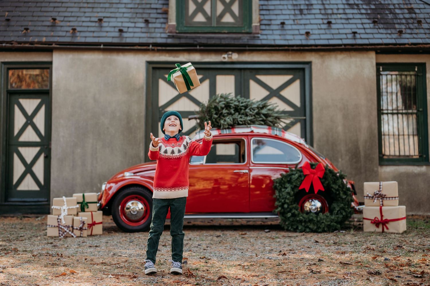 A vintage red Volkswagen Beetle decorated with Christmas wreaths and presents sits outside a rustic barn.