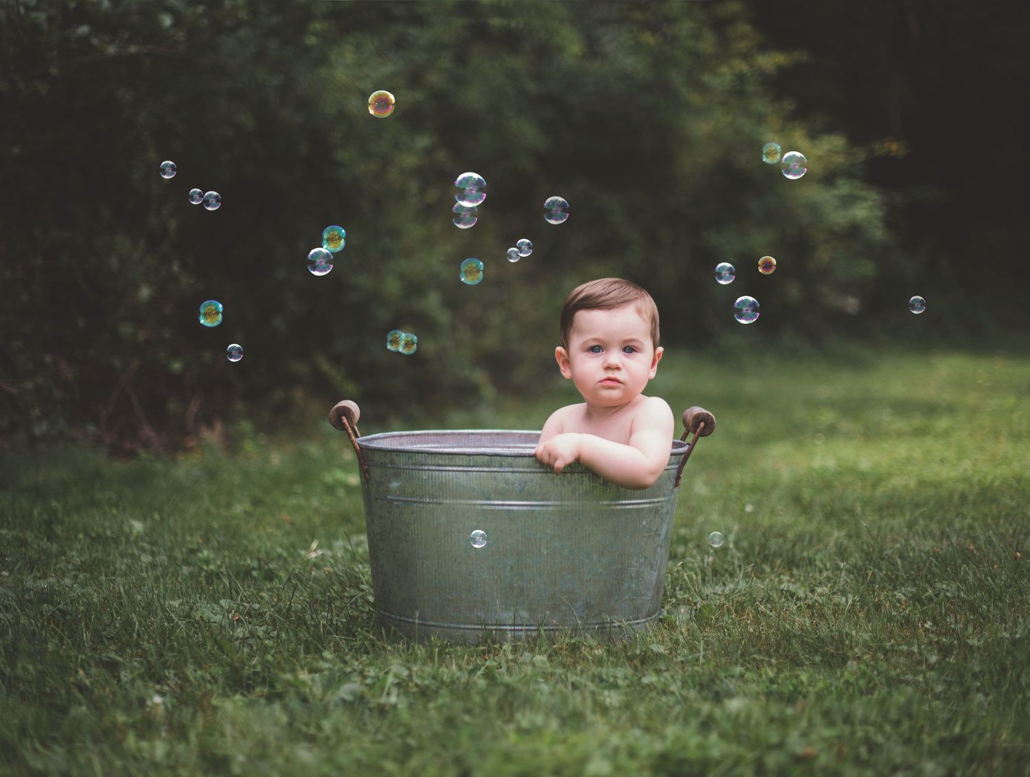 A small child bathes in a vintage metal tub outdoors while soap bubbles float through the air against a green background.