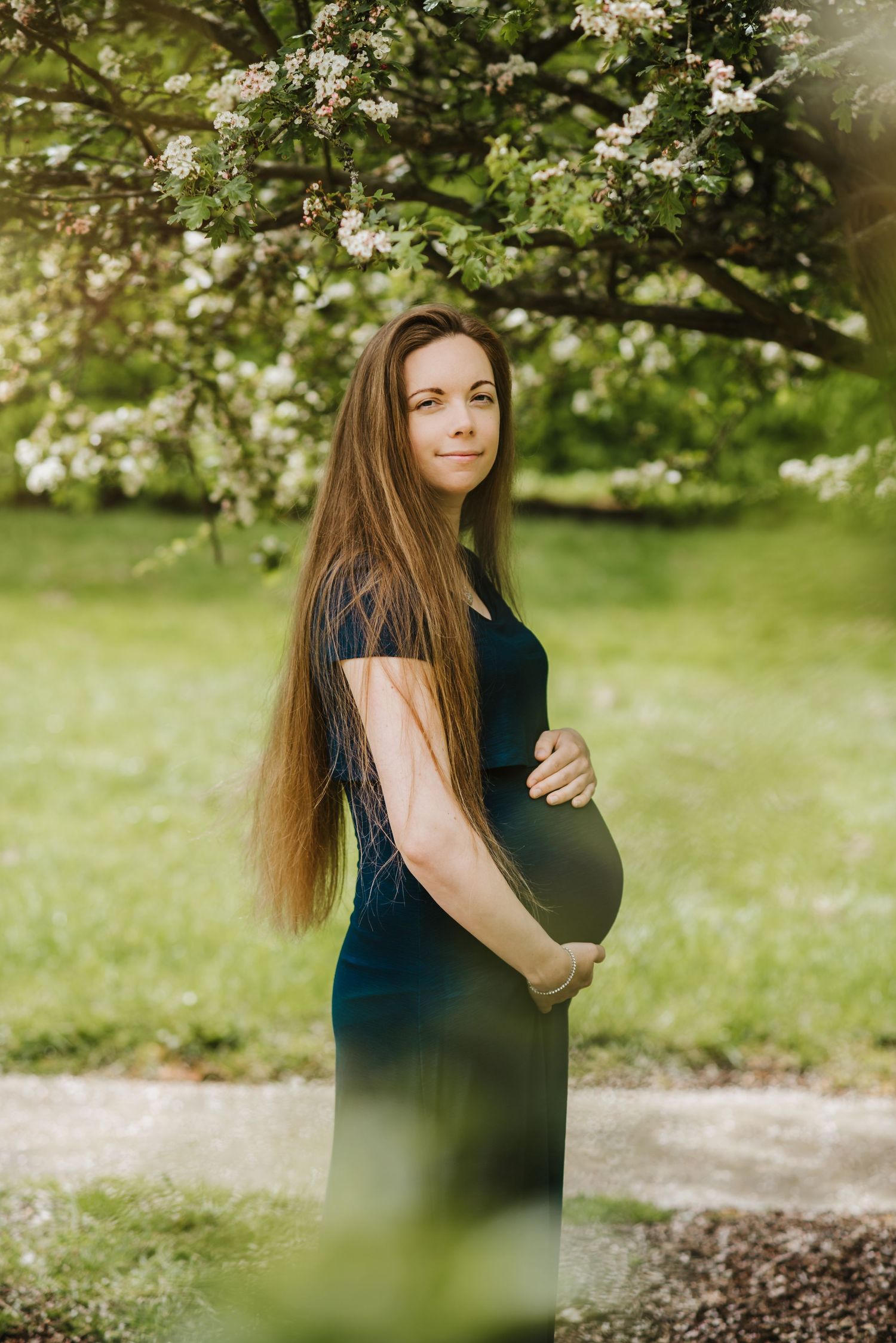 Maternity photo in a garden with blooming trees and soft sunlight filtering through spring blossoms.