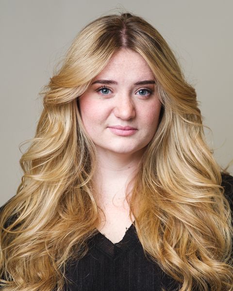 Studio portrait of a person with long wavy blonde hair wearing a black v-neck top against a neutral background.