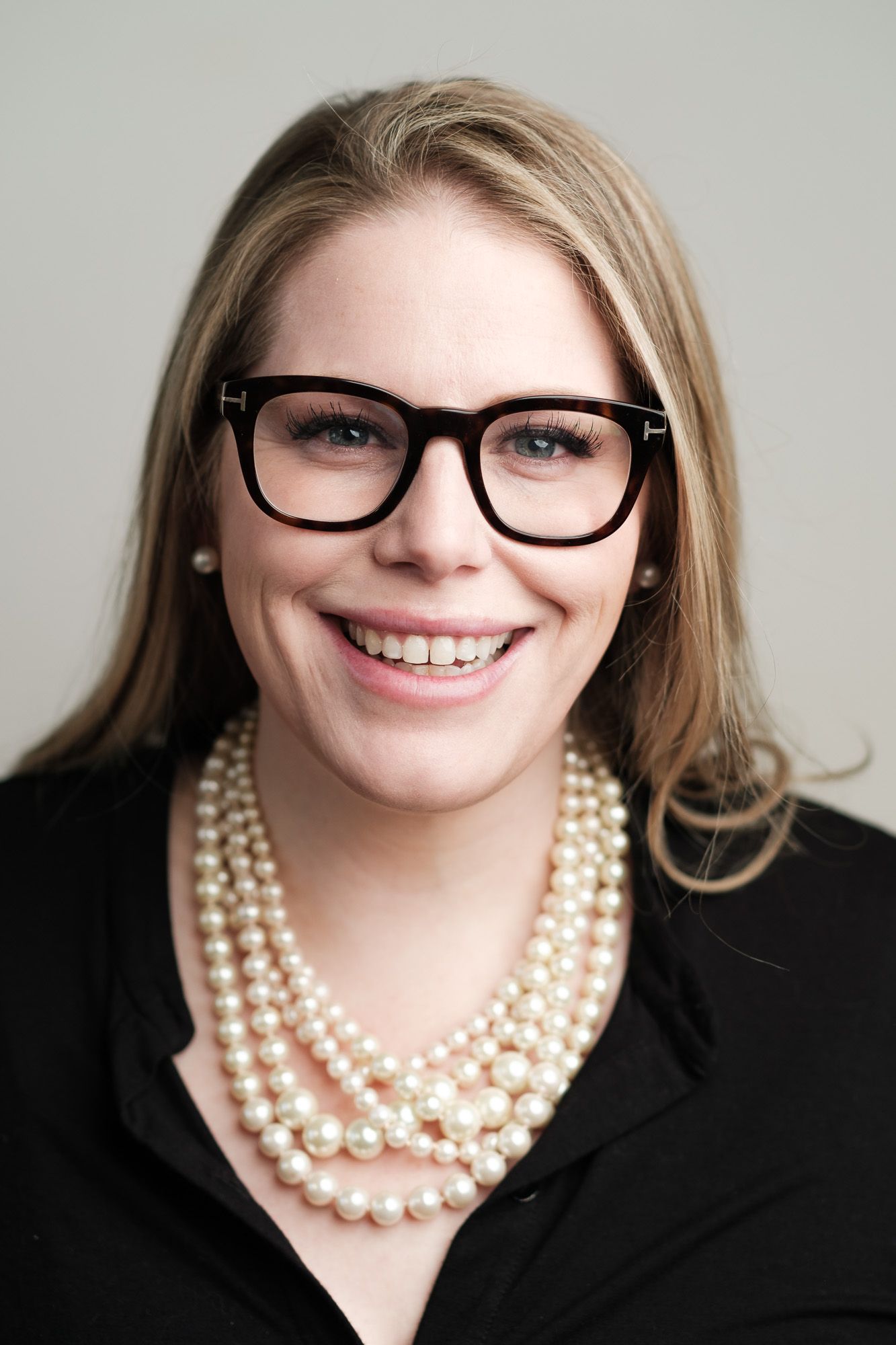 Professional headshot with black-rimmed glasses and layered pearl necklace against light background.