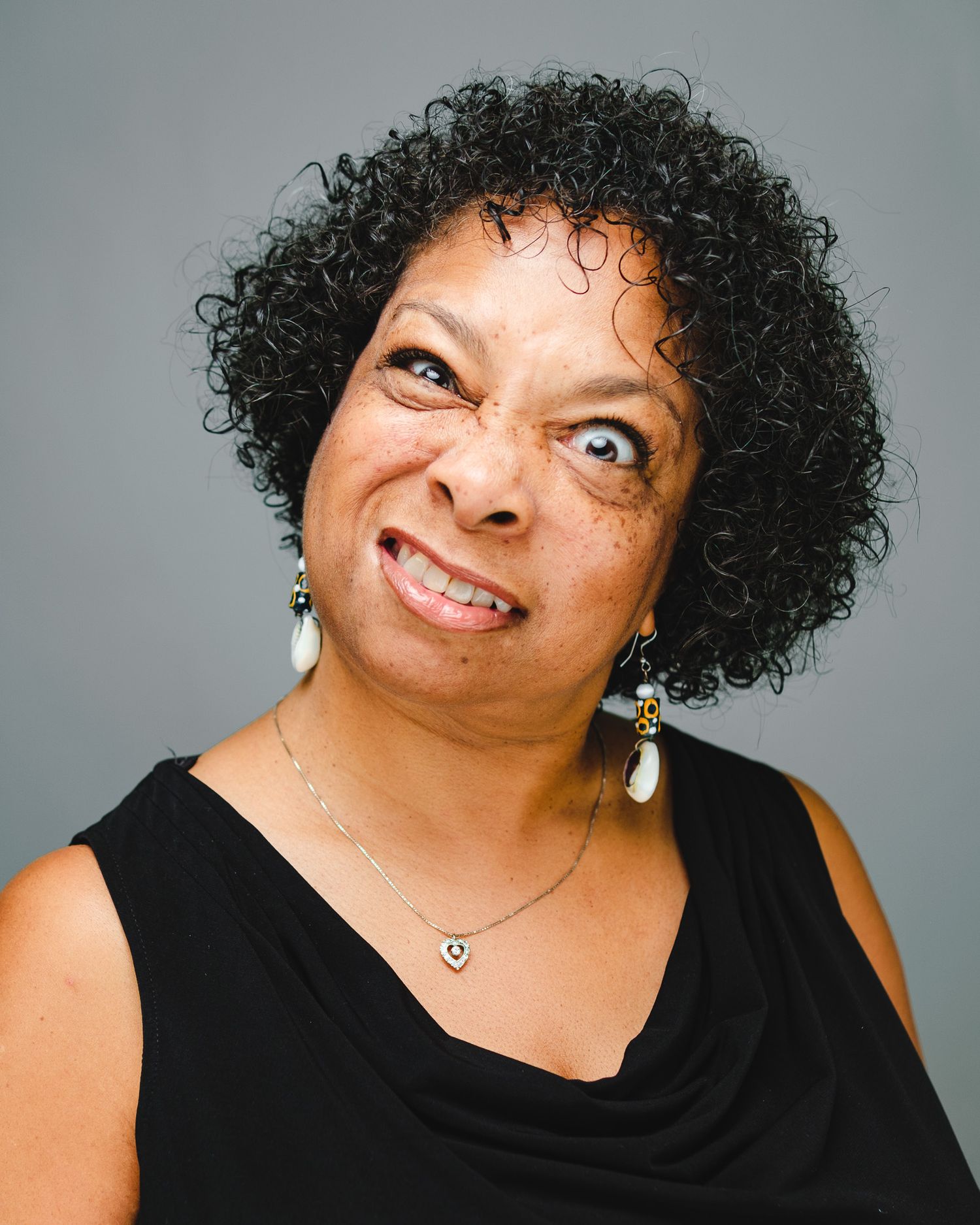 A funny professional headshot showing a crazy smile against a gray background, wearing a black top and teardrop earrings.
