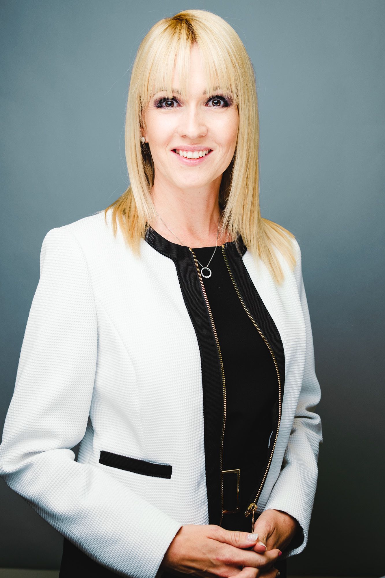 Professional portrait of a professional woman in a crisp white blazer and black top against a gray background, featuring blonde hair.