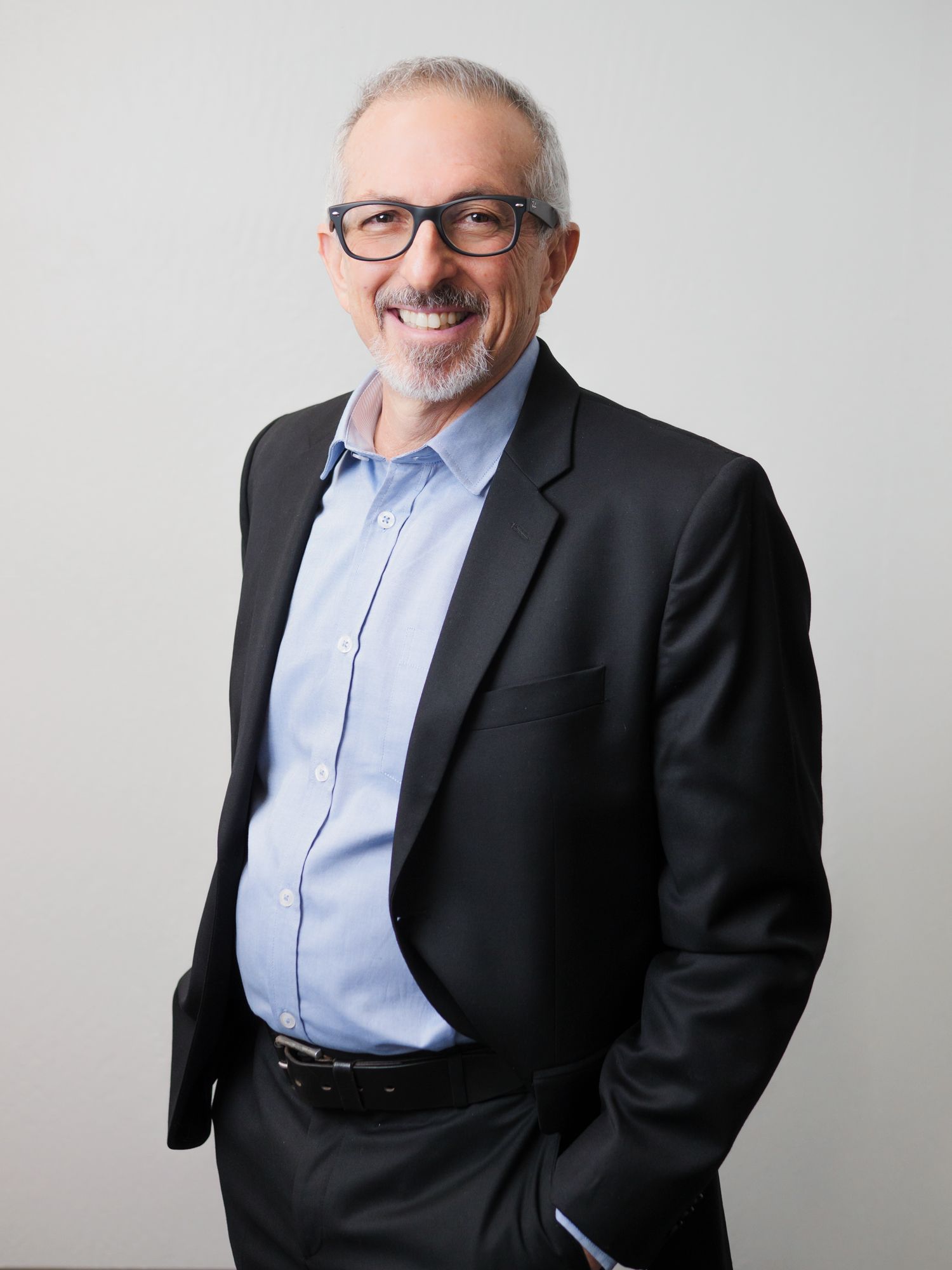 Professional business portrait of someone wearing glasses, a light blue dress shirt and dark suit jacket against gray background.