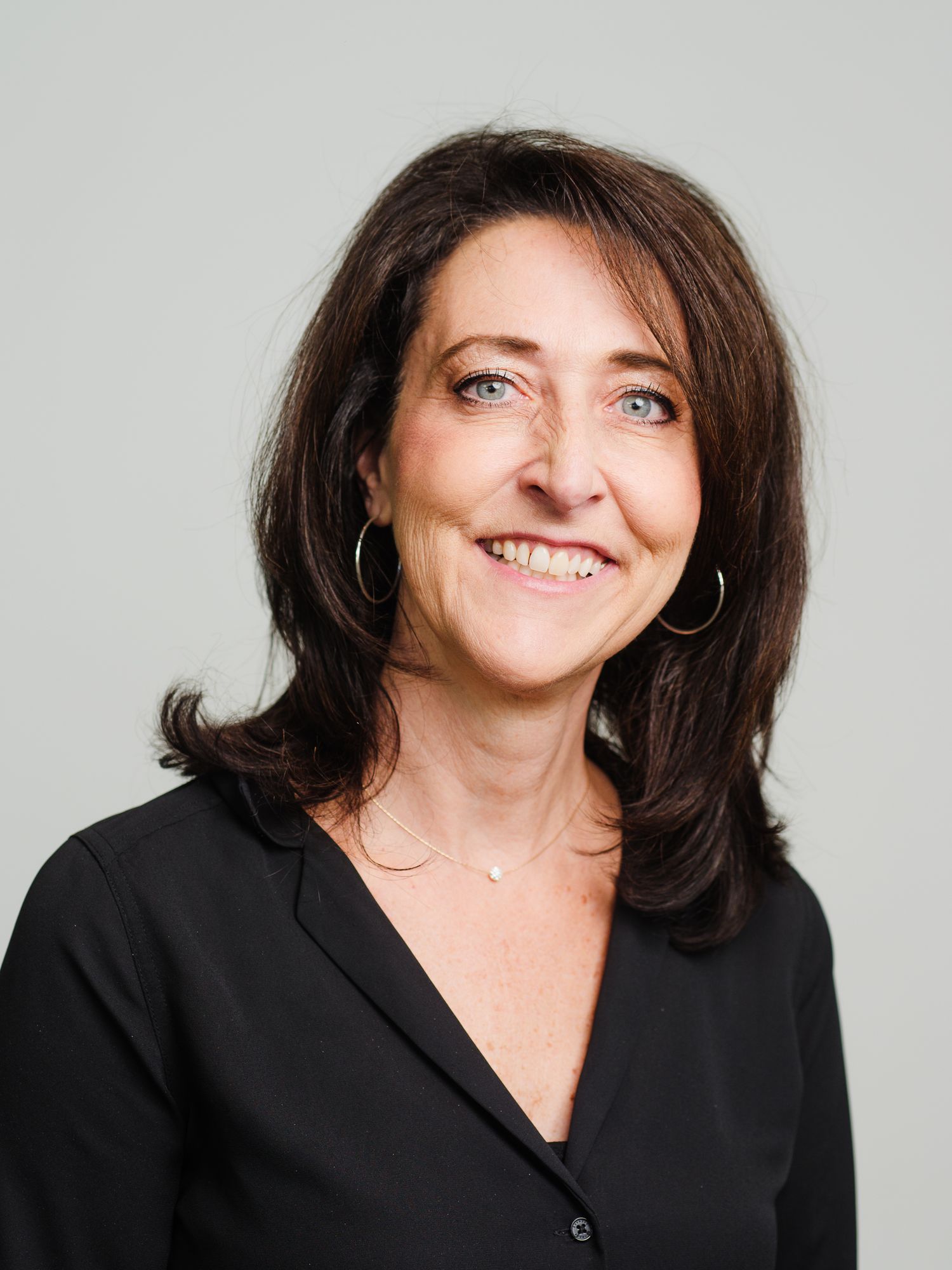 Professional headshot portrait of someone with dark hair wearing black blazer and silver hoop earrings against light background.