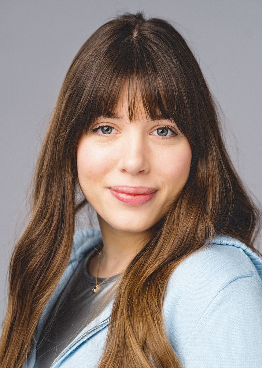 Professional headshot portrait of someone with long brown hair and bangs wearing a light denim sweater against neutral background.