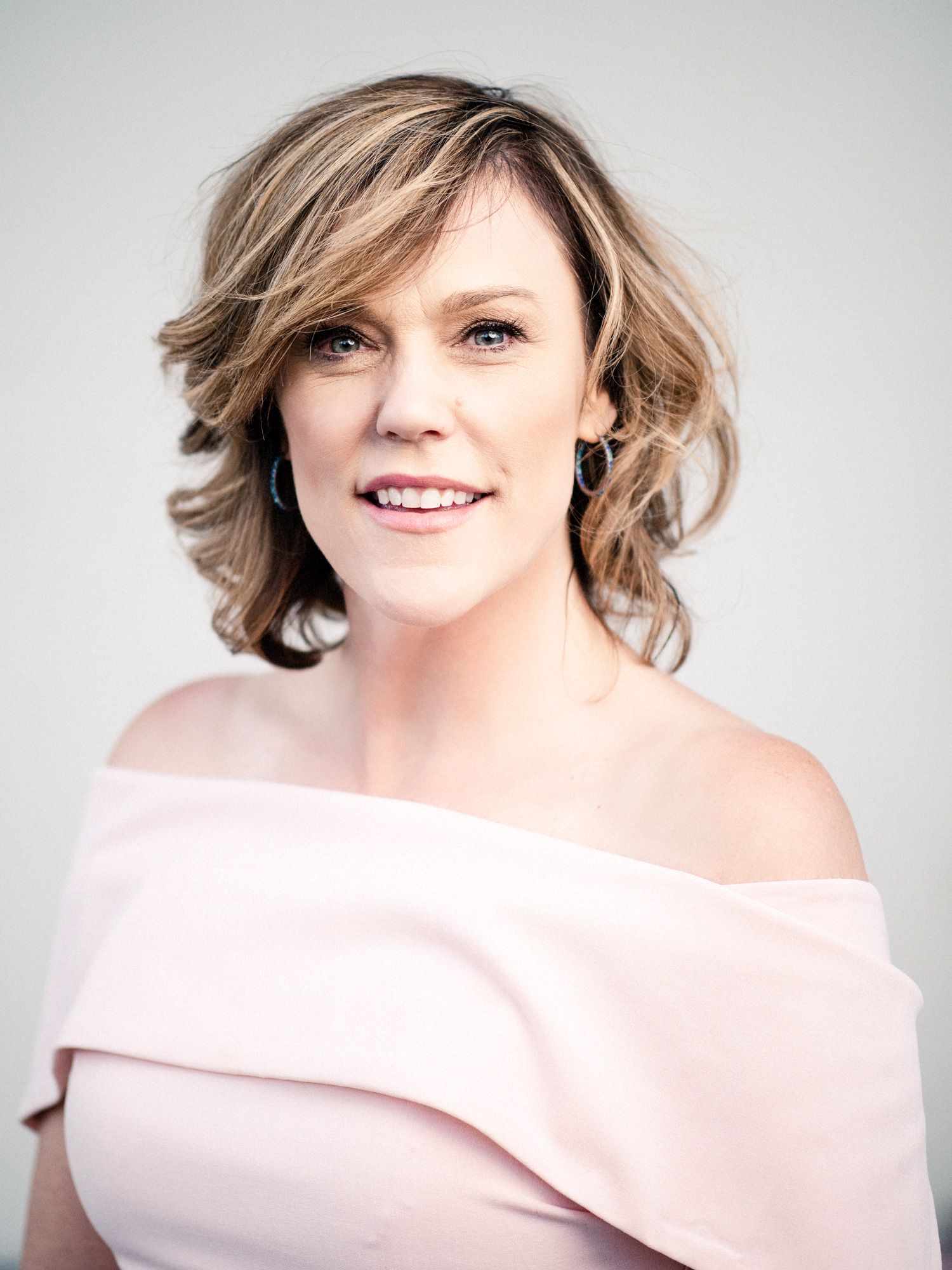 Professional portrait showing a warm smile and wavy blonde hair, wearing an elegant off-shoulder pink dress against a light background.