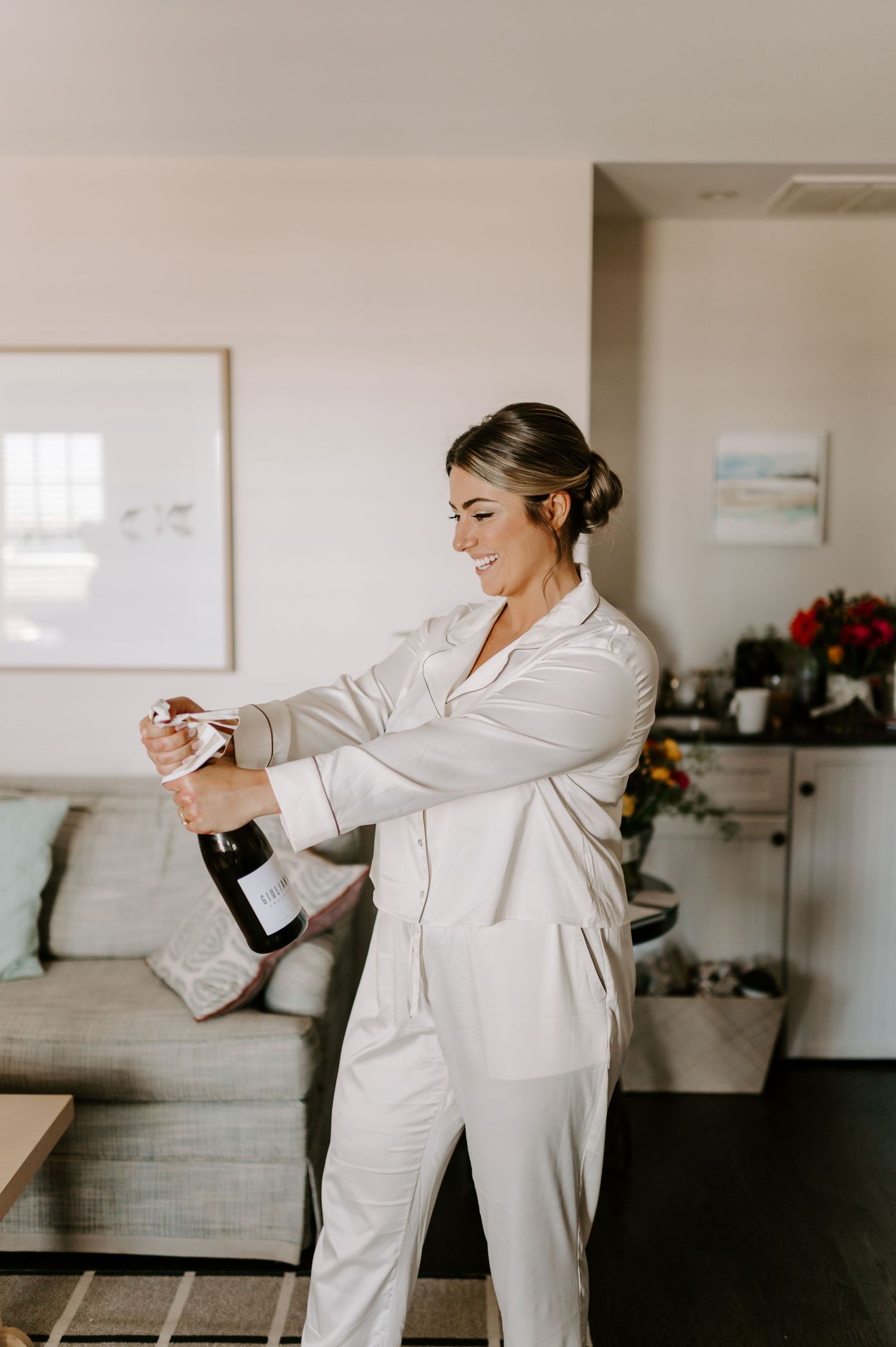 Person in white robe opens champagne bottle during wedding preparations.
