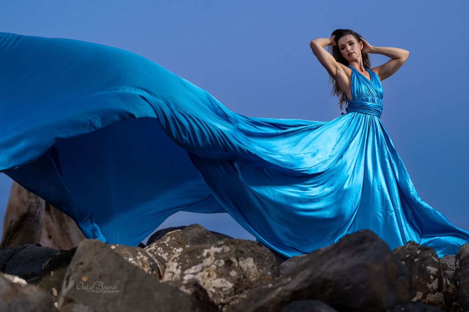 Model in flowing blue gown creates dramatic silhouette against rocky landscape.