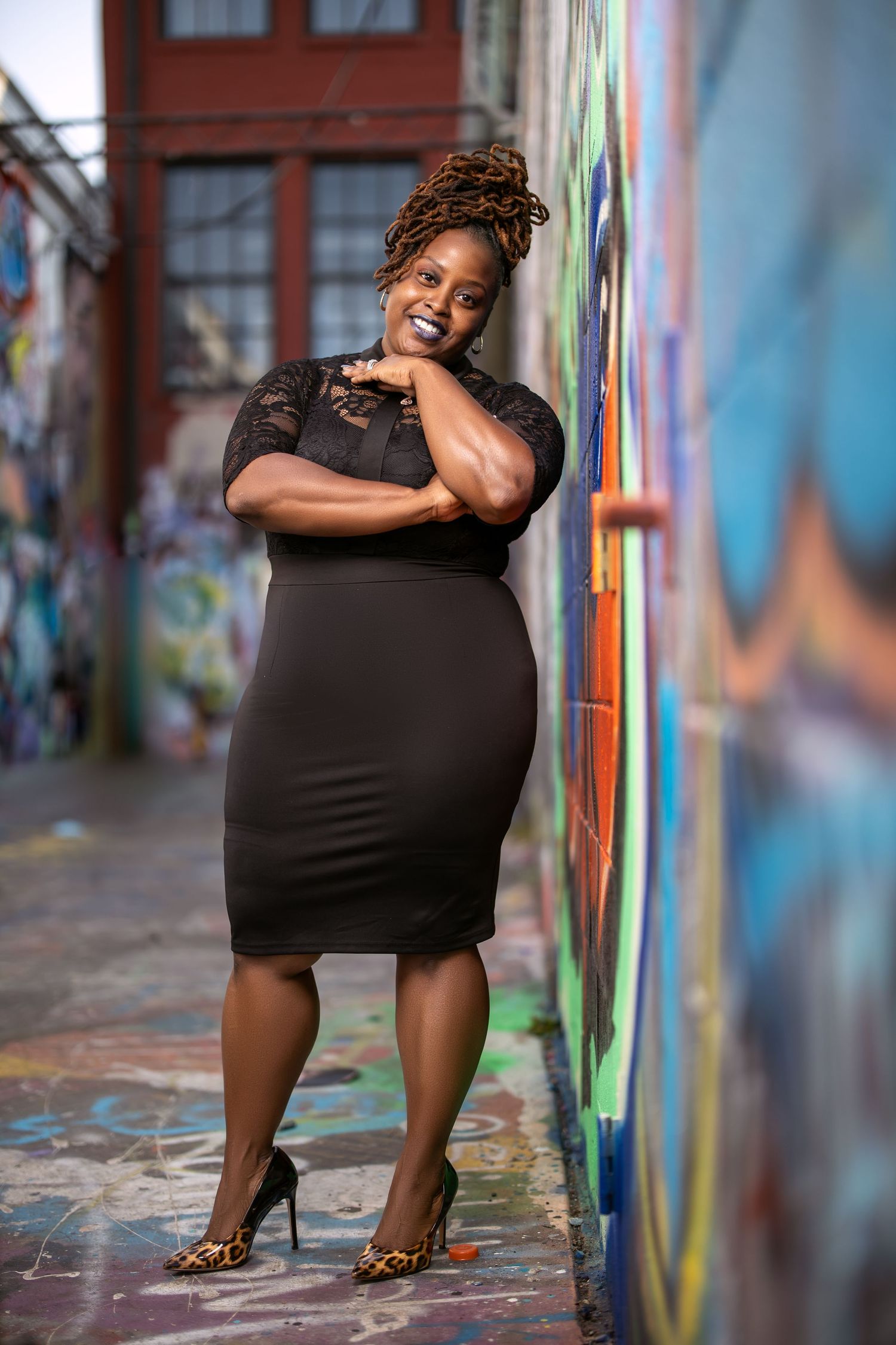 Woman in black dress poses stylishly against vibrant street art wall in urban environment.