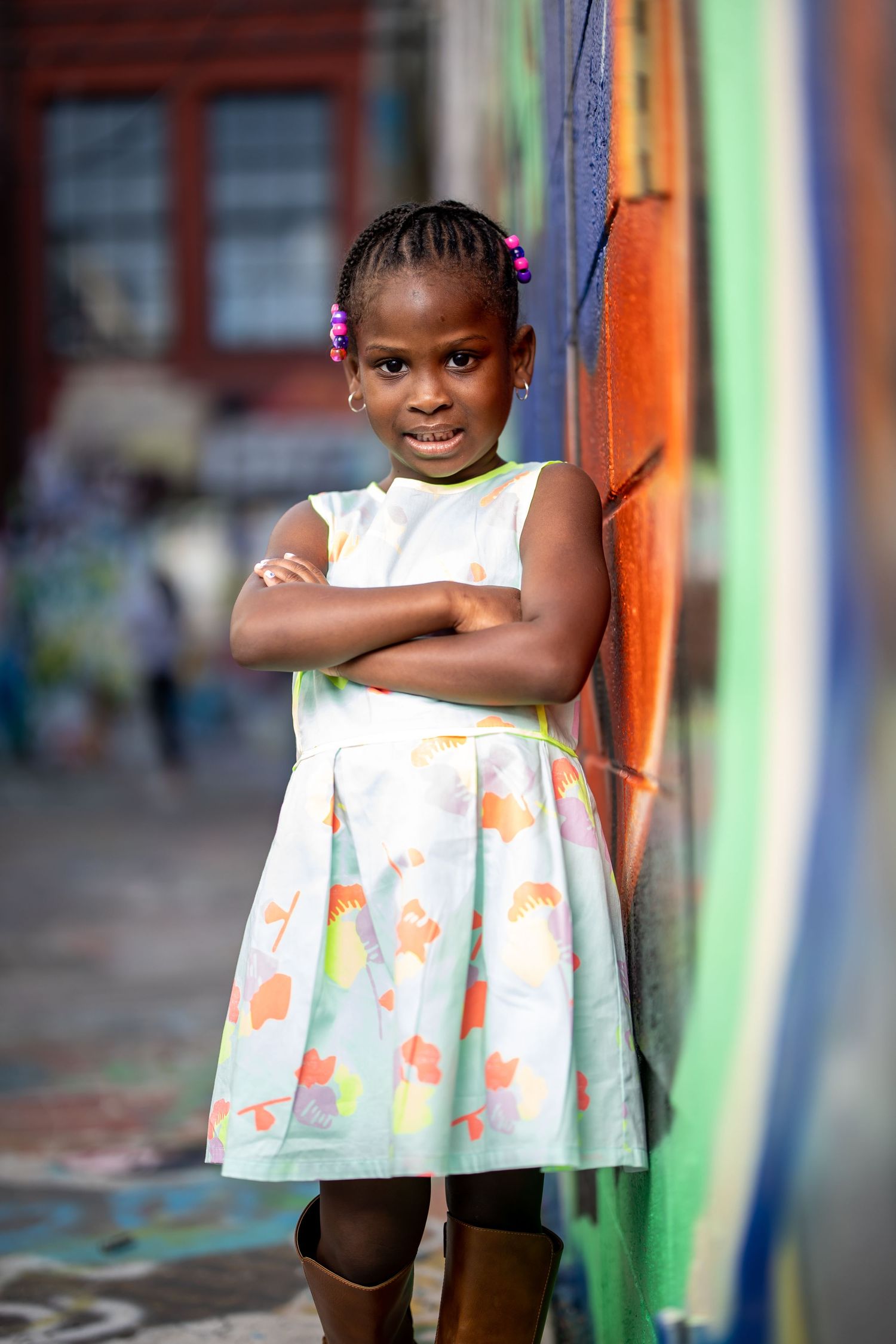 Young child in a floral white dress poses confidently against a colorful graffiti wall in an urban setting.